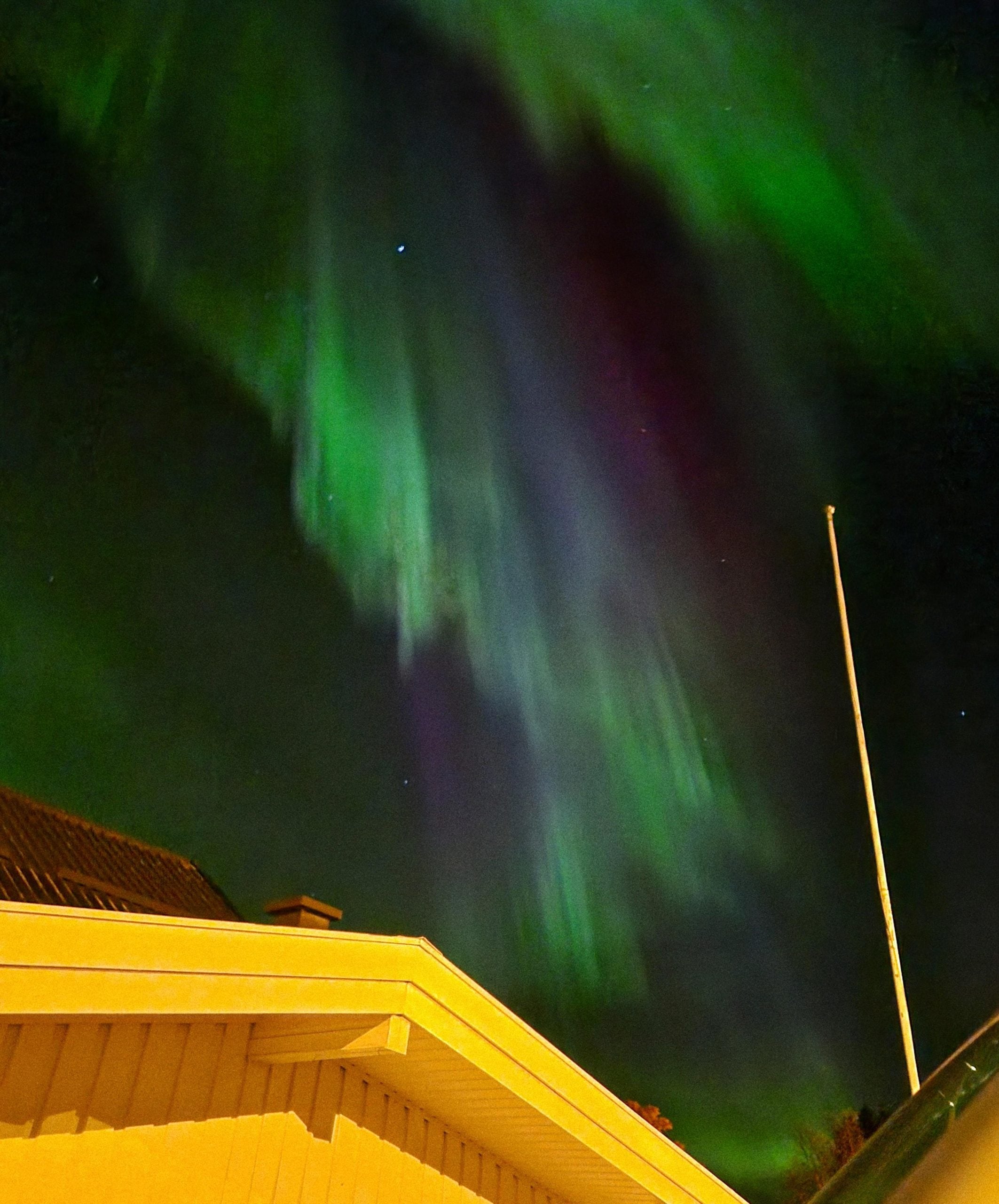 Green and purple aurora borealis in the night sky above a house roof and flagpole.