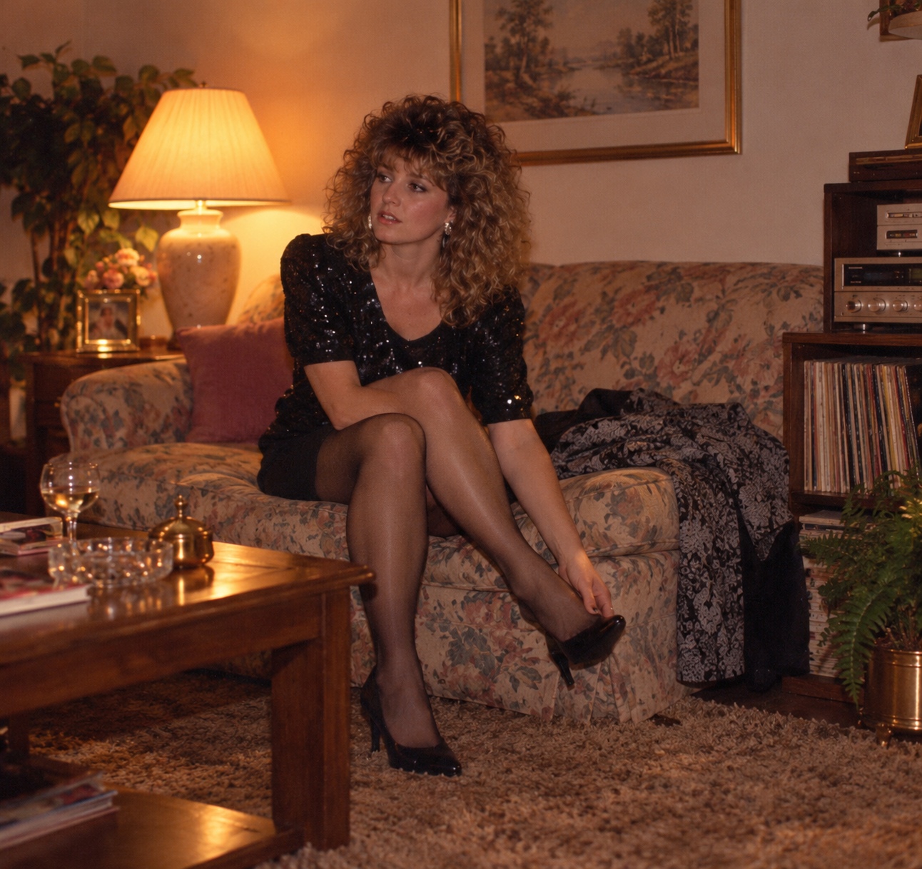 A woman with curly hair wearing a sequined top and stockings sits on a floral sofa while putting on a black high-heeled shoe.