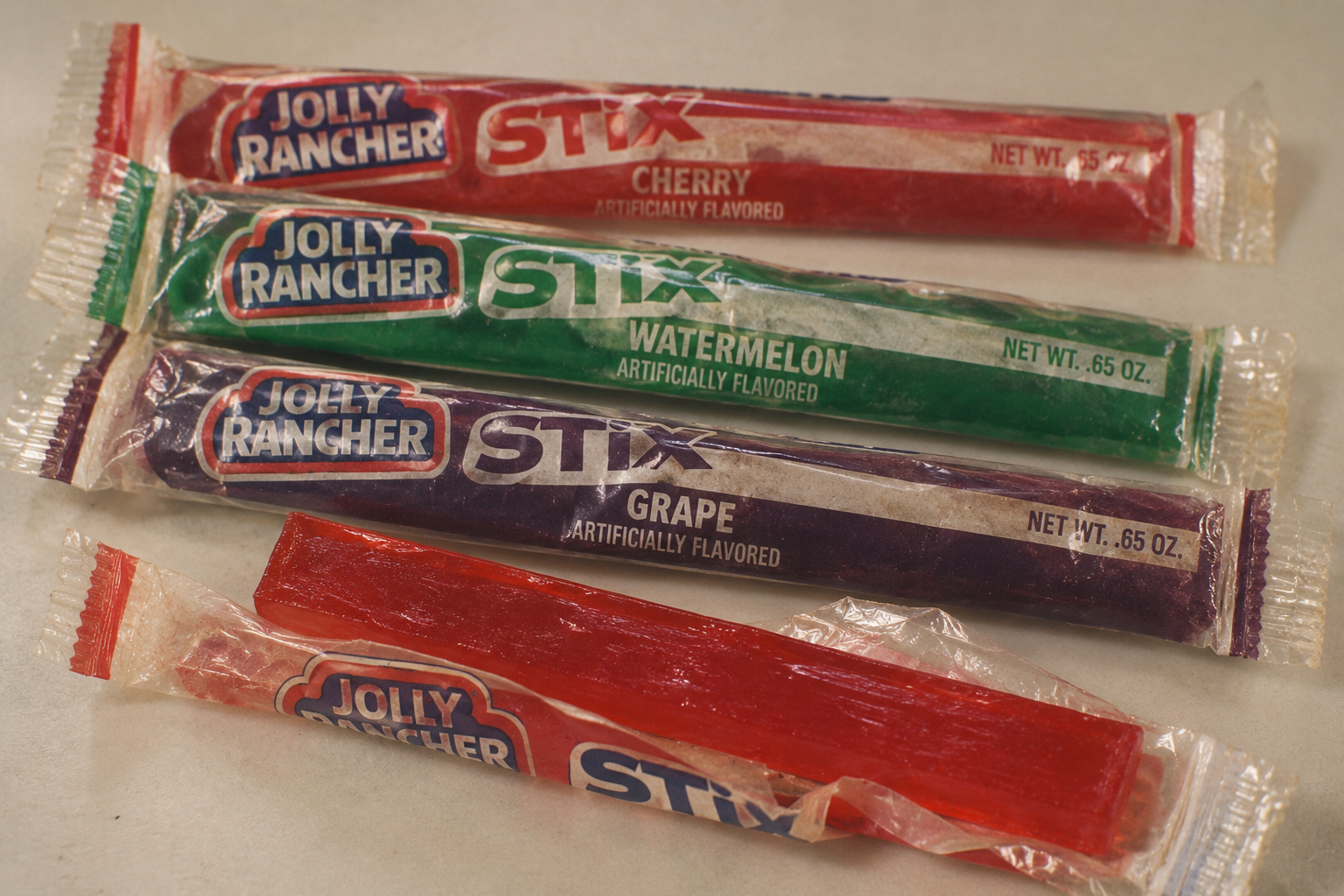 Three wrapped Jolly Rancher Stix candy bars in cherry, watermelon, and grape flavors, with one unwrapped red cherry candy bar in the foreground.