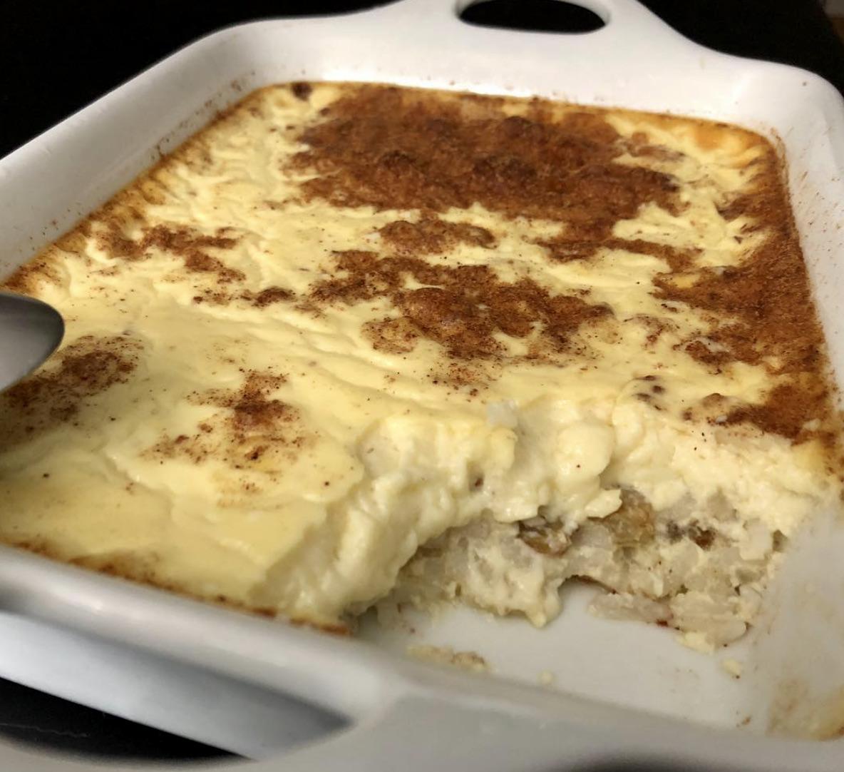 A white baking dish contains creamy rice pudding topped with cinnamon, with a spoon and a portion already served, revealing the textured inside.