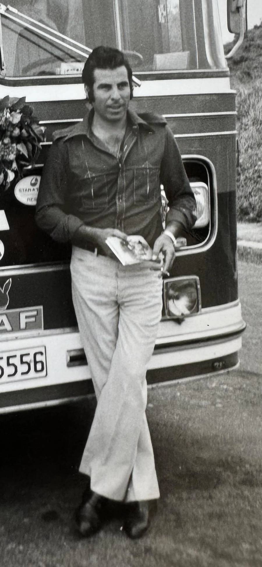 A man standing with his legs crossed, leaning against the front of a bus while holding a small booklet.