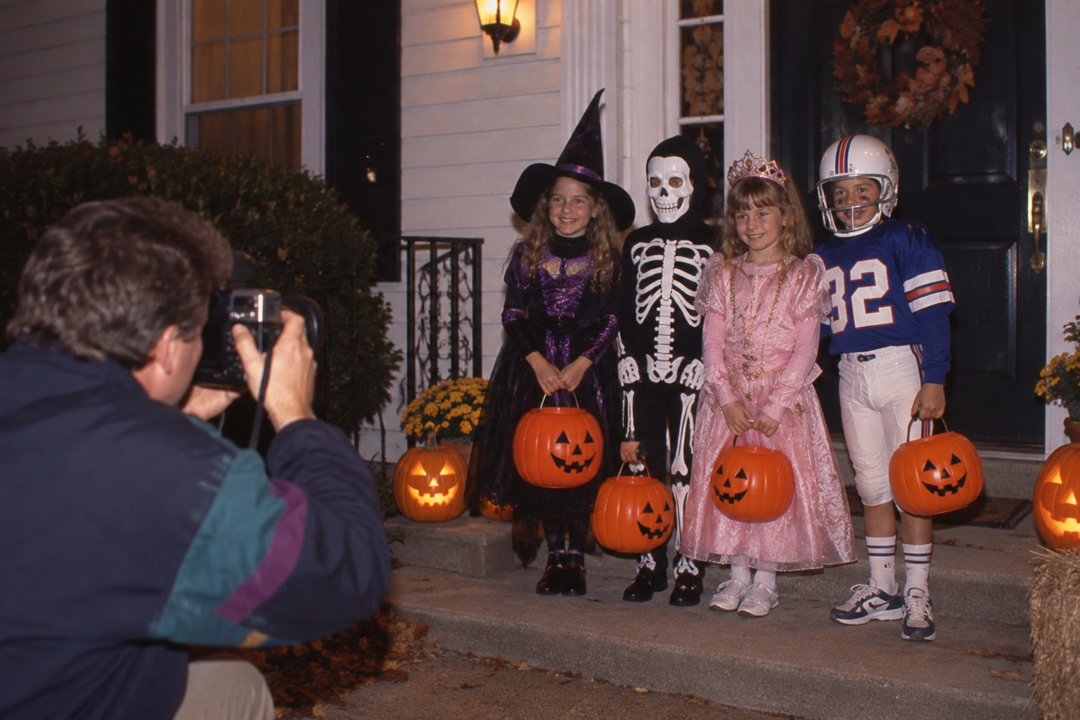 A photographer takes a picture of four children in Halloween costumes standing on a front porch with jack-o’-lanterns.