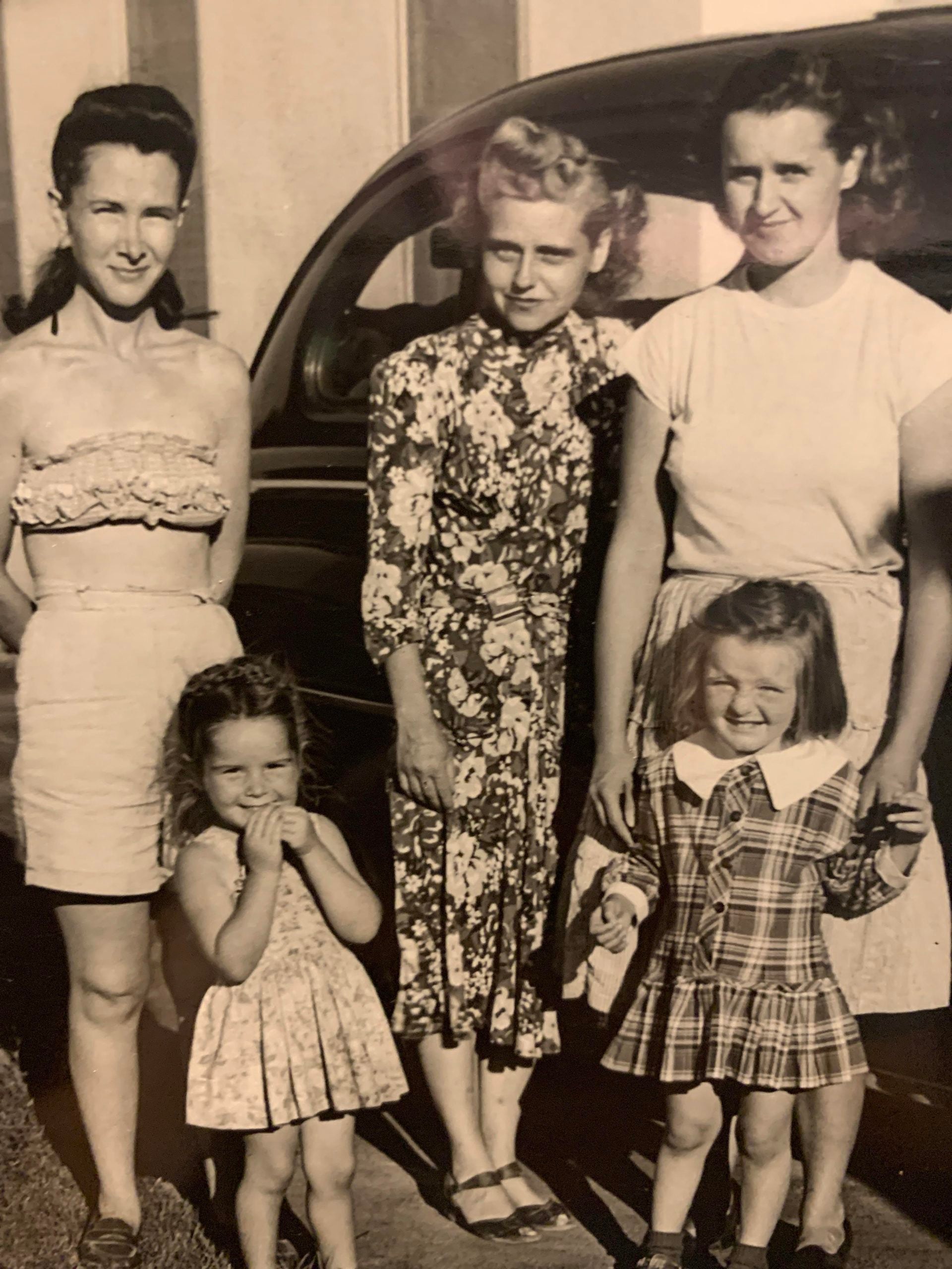 Three women and two young girls posing for a vintage photograph in front of a car.