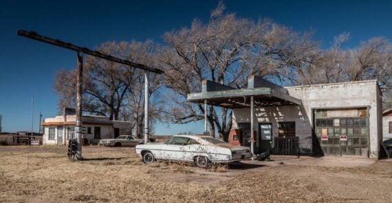 A vintage white car sits parked under the canopy of a derelict gas station in a dry, open field.