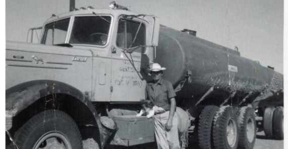 A man in a cowboy hat stands next to a tanker truck holding a small dog.