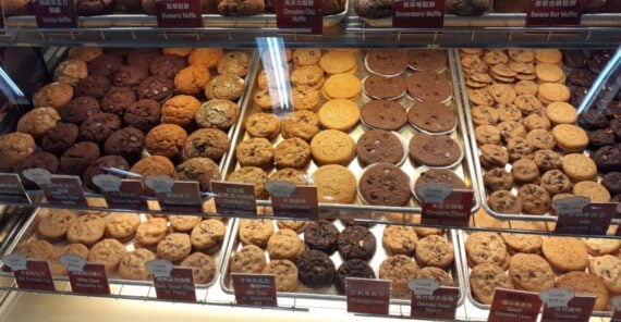 A glass display case filled with rows of assorted muffins and cookies, each with a small label indicating its flavor.