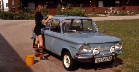 A person in shorts and striped socks washes a light blue NSU Prinz car parked in front of a house.
