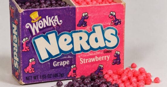A box of Wonka Nerds candy with purple grape and pink strawberry compartments, with loose candy of both flavors spilled in front.
