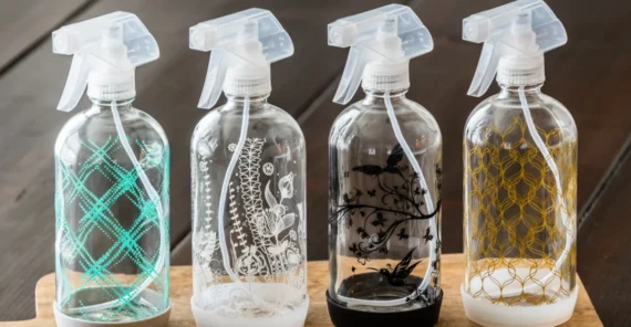 Four decorative glass spray bottles with different etched patterns sit on a wooden tray atop a dark wooden table, with a small green potted plant blurred in the background.