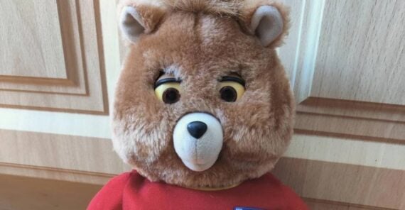 A close-up of a vintage Teddy Ruxpin plush bear with a light brown face, round ears, expressive eyes, and a red shirt, seated in front of wooden paneling.