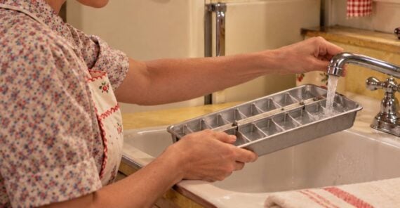 A woman in an apron fills an empty metal ice cube tray with water at a kitchen sink.