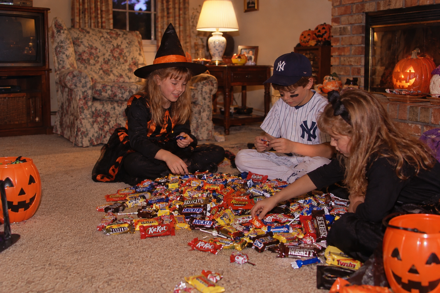 Three children in costumes sit on a living room floor sorting through a large pile of assorted Halloween candy.