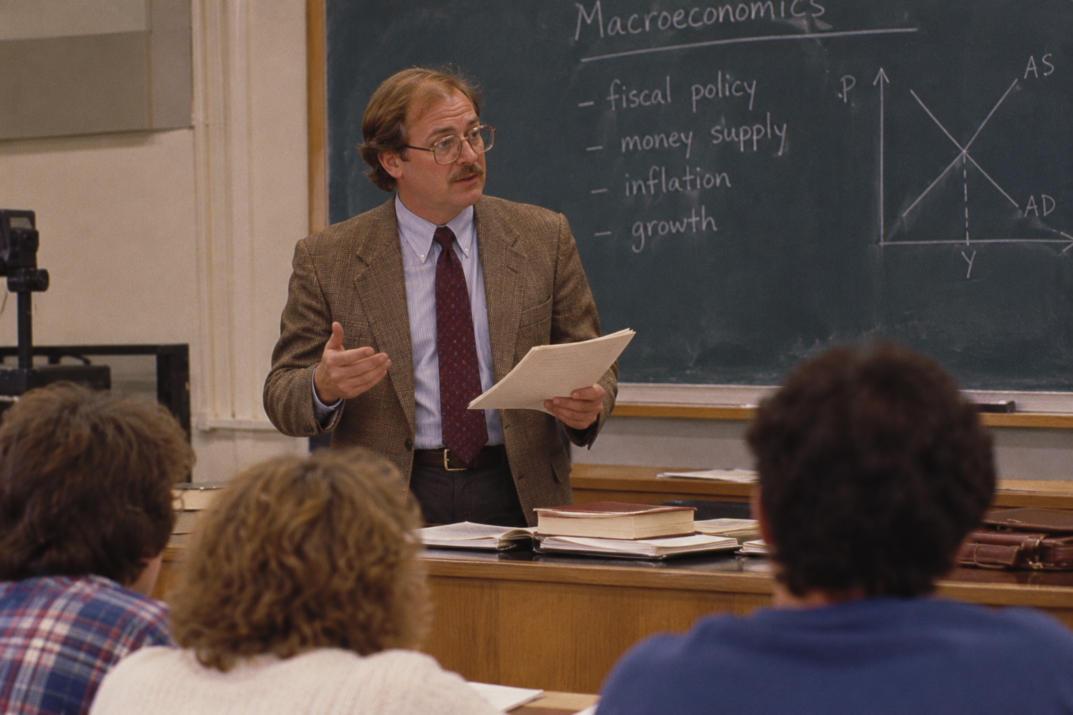 A professor in a blazer stands before a blackboard lecturing a class, gesturing with notes in his hand while a macroeconomics graph and list of topics are written behind him.