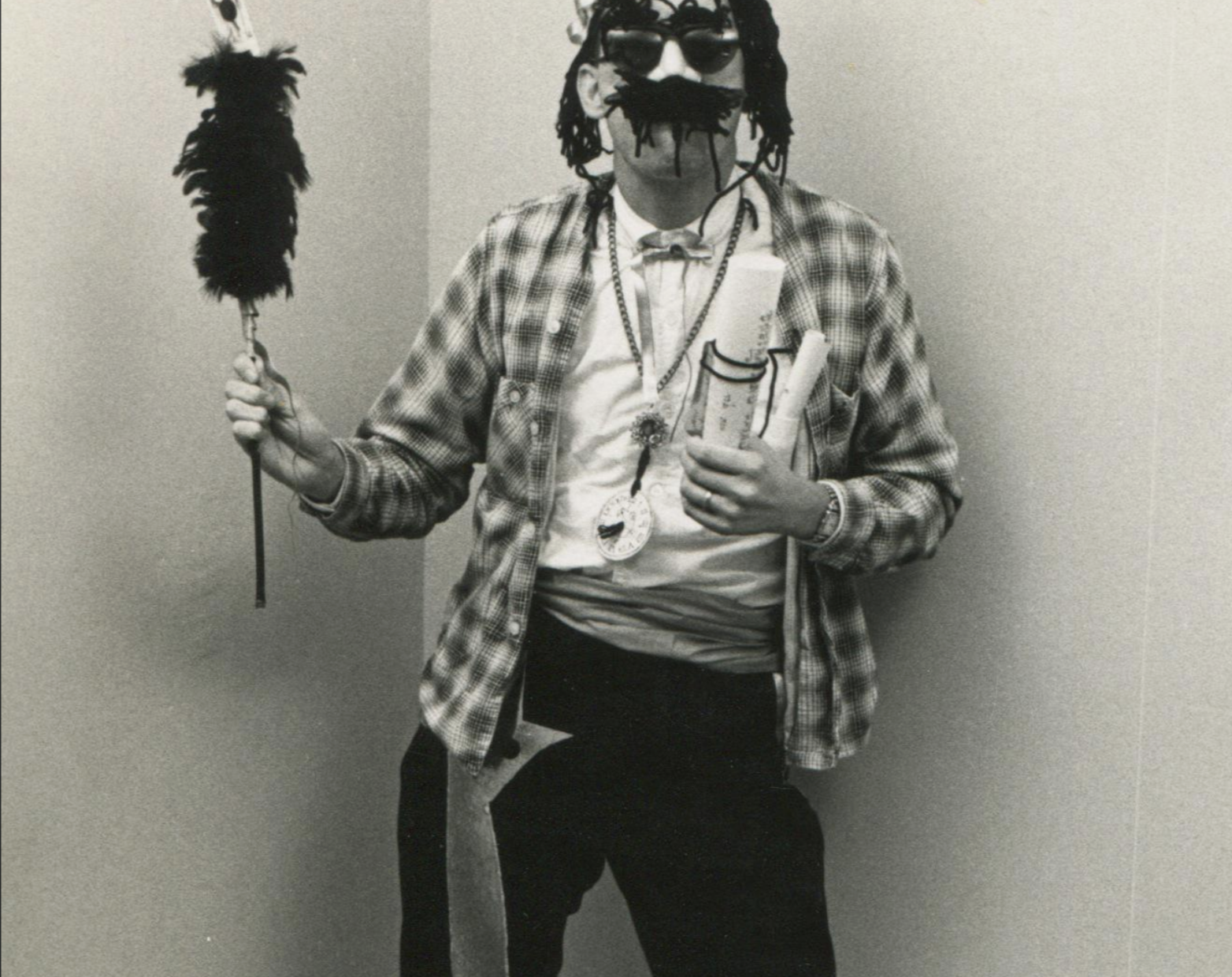 A person in a plaid shirt, sunglasses, and a fake mustache holds a feather duster and a rolled document.