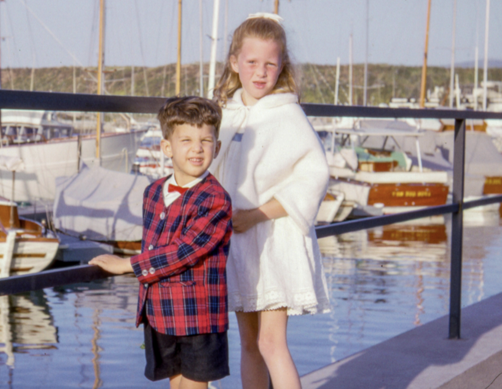 A young boy in a plaid blazer and red bowtie stands next to a young girl in a white dress and cape on a dock in front of a marina.
