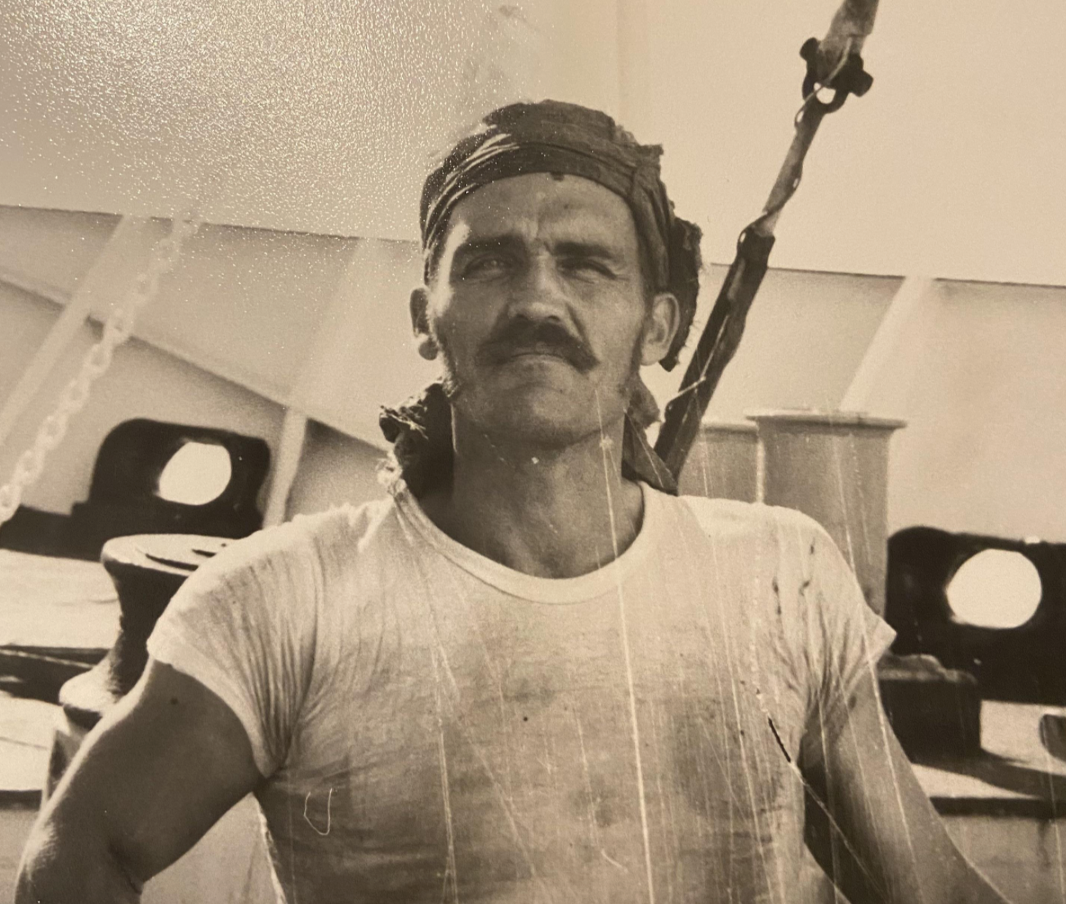 A black-and-white portrait of a man with a mustache wearing a headscarf on the deck of a ship.