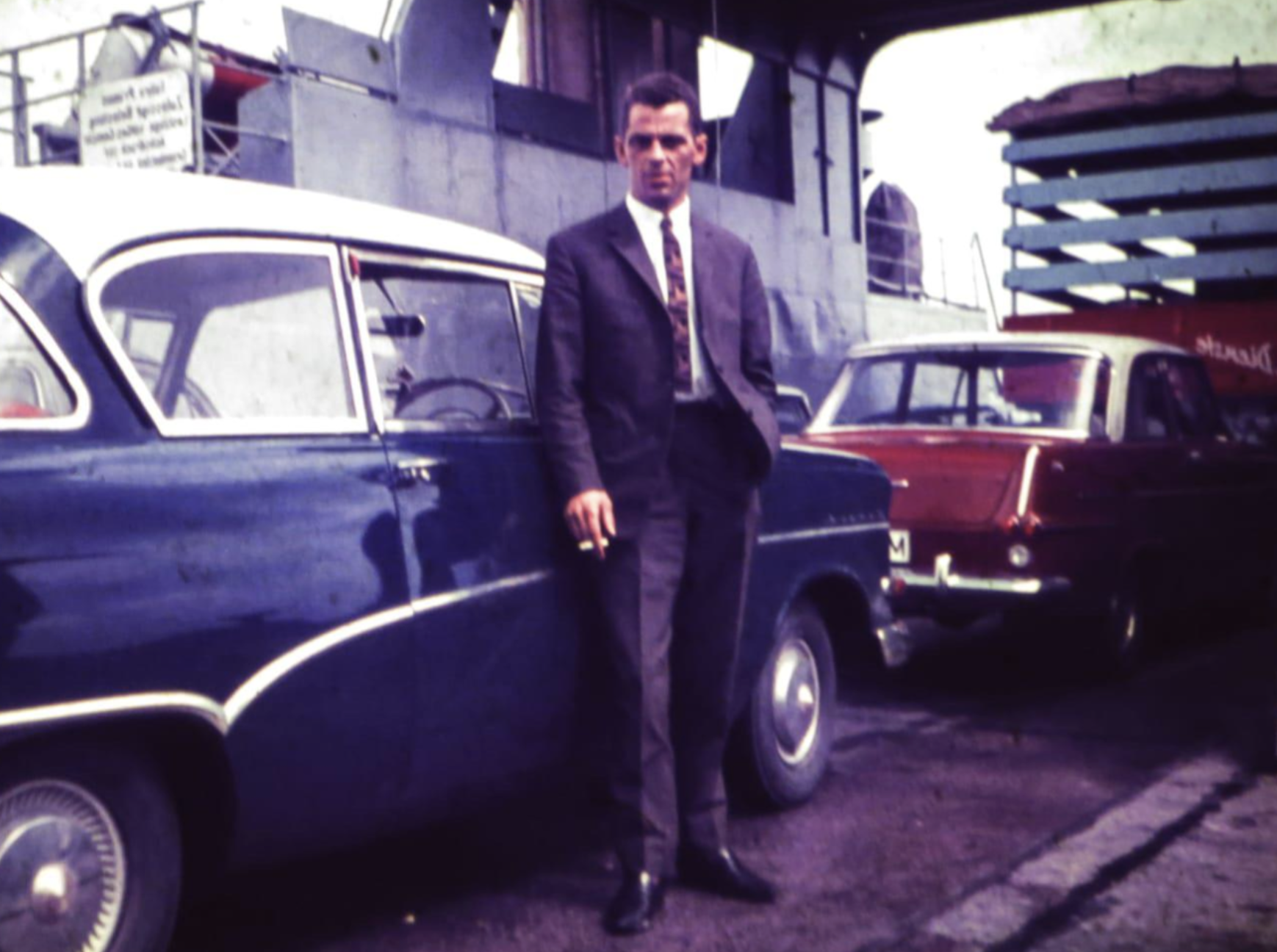 A man in a suit stands leaning against a parked dark blue car on a ferry deck.