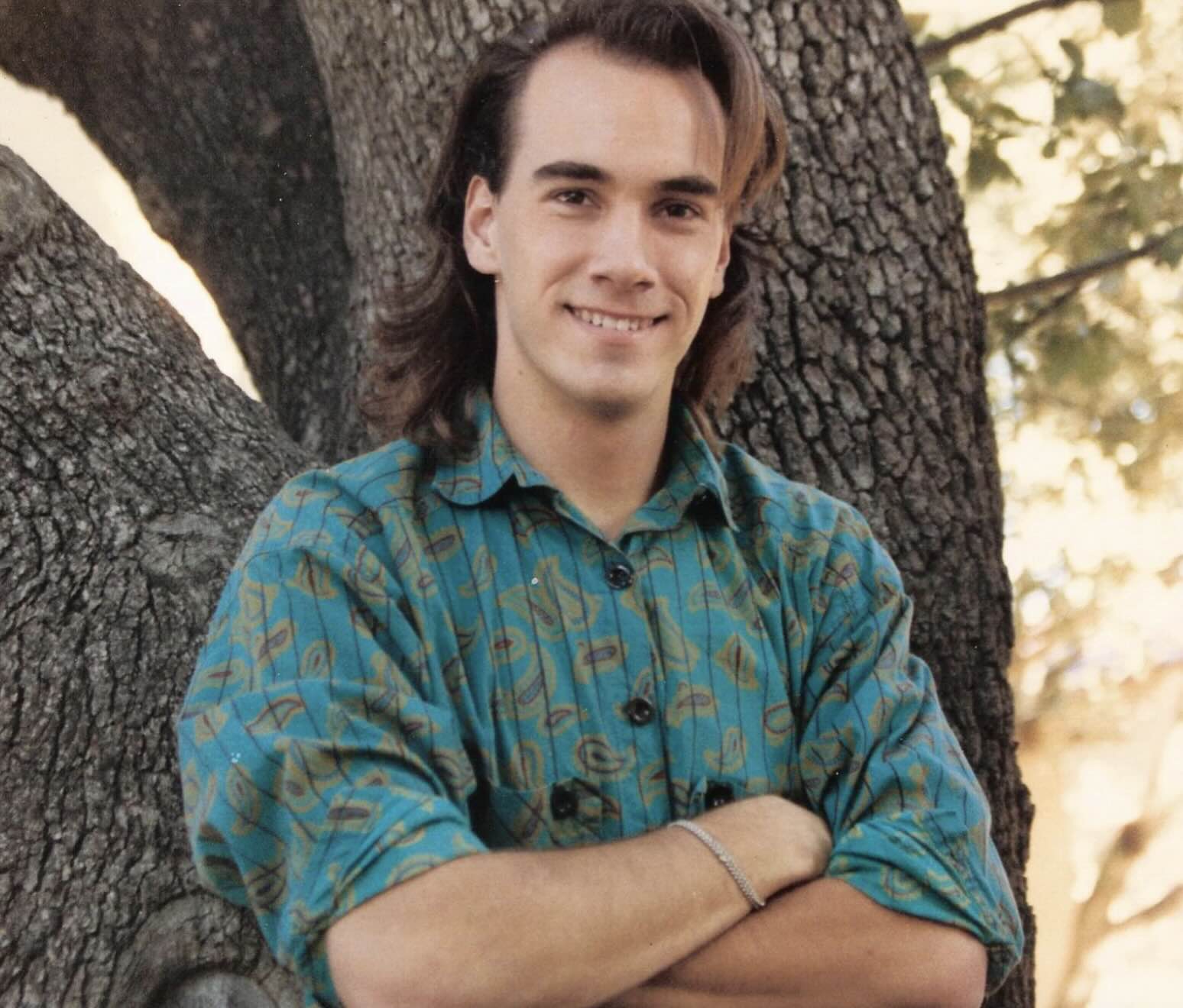 A young man with shoulder-length hair and a patterned teal shirt stands with his arms crossed in front of a tree trunk.