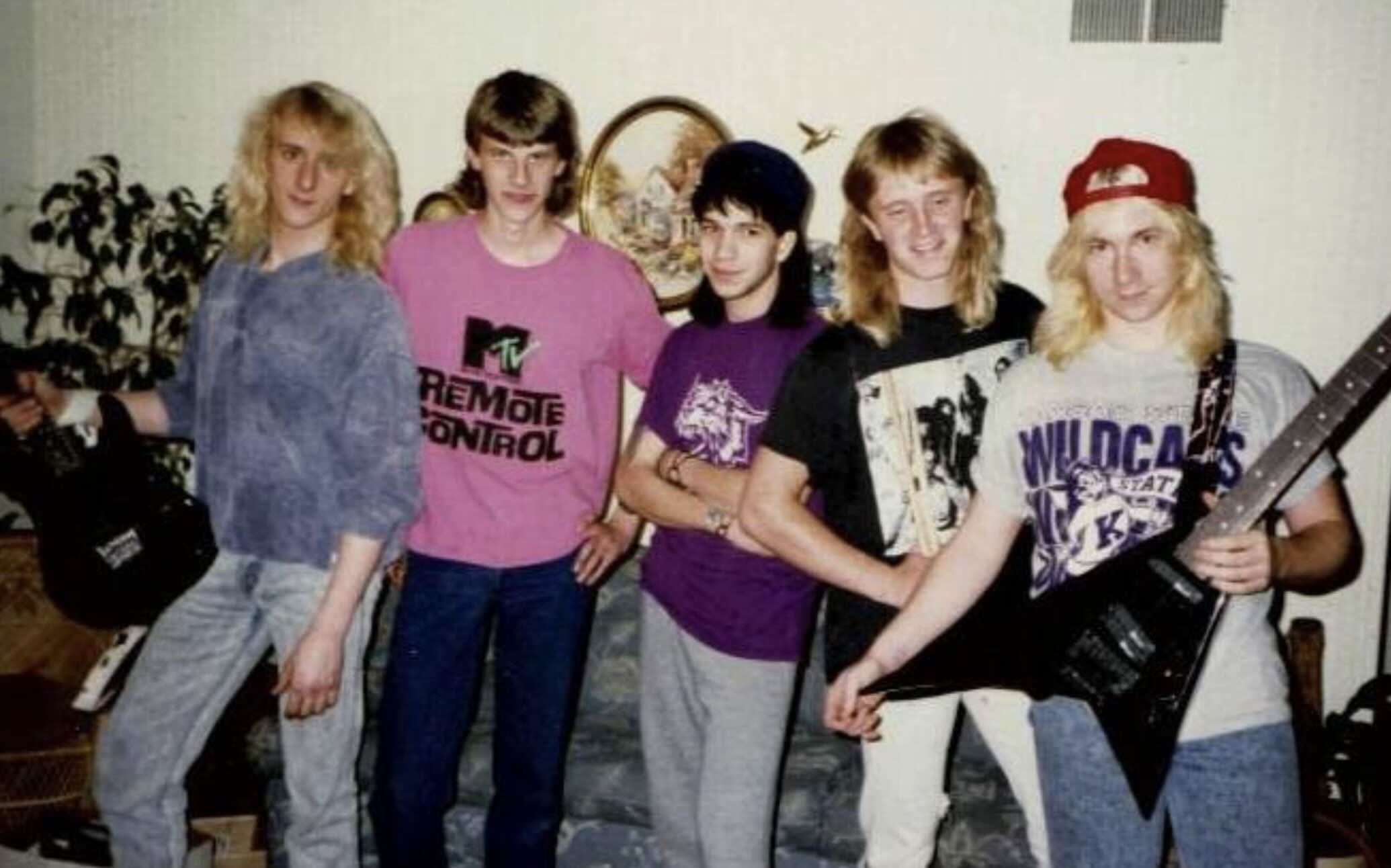 Five young men with long hair posing together in a room, two of whom are holding electric guitars.