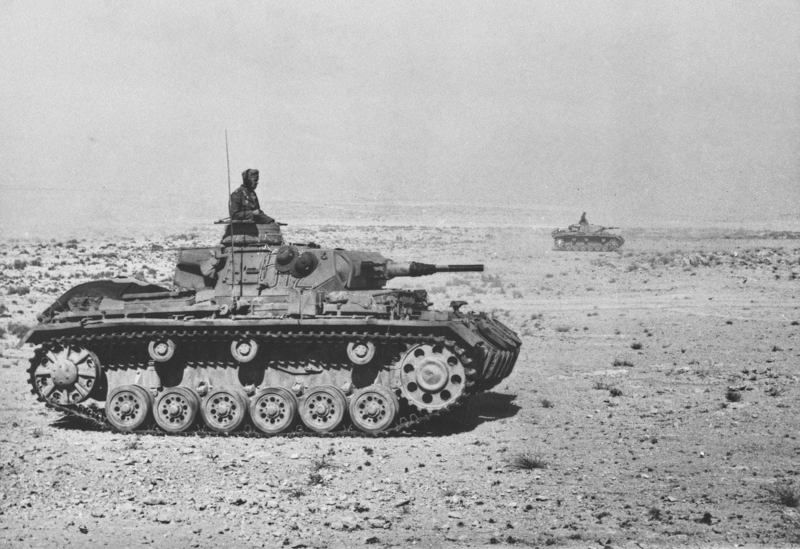 A World War II-era tank with a soldier on top moves across a rocky desert landscape; another tank is visible in the distance under a hazy sky.