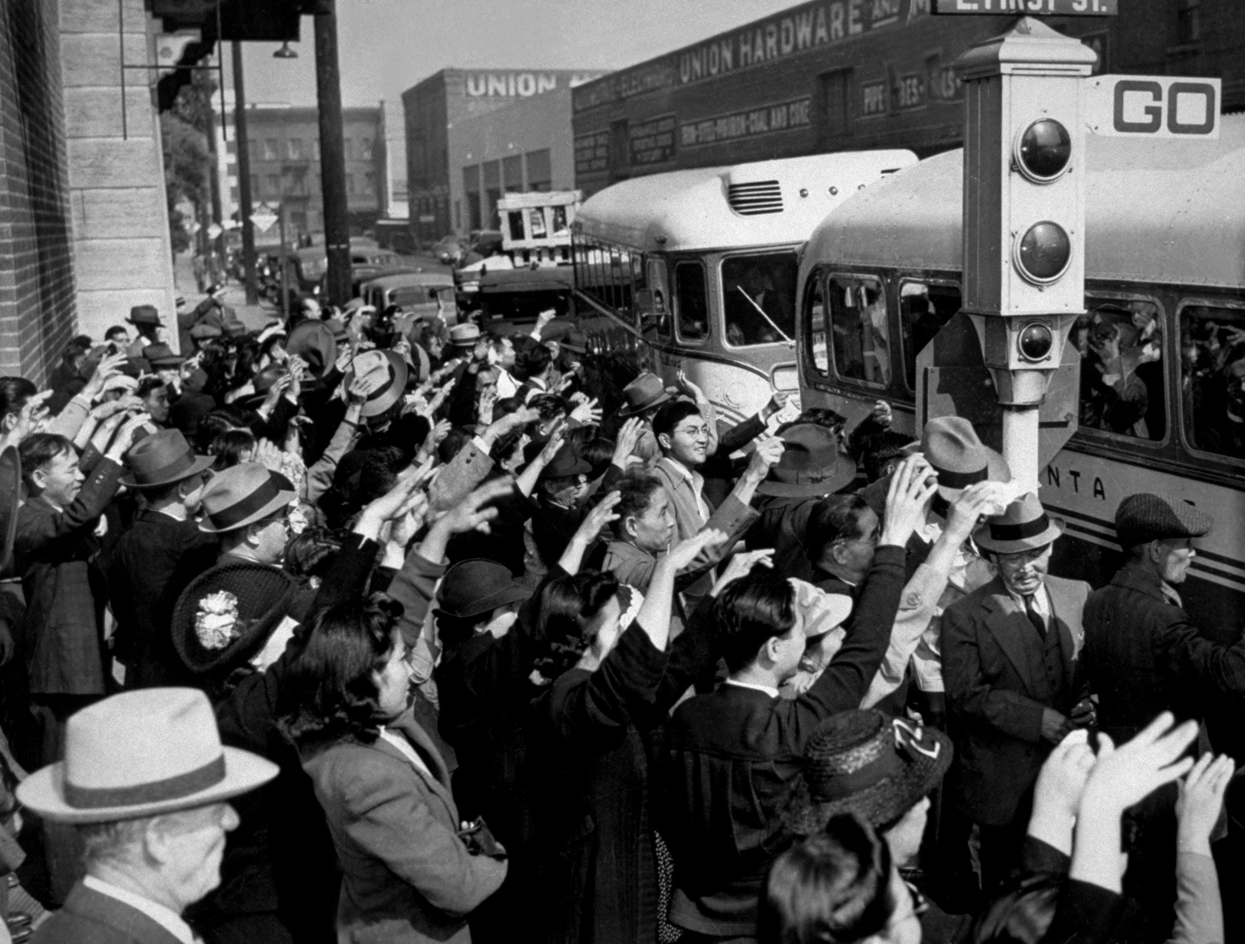 A crowd of people waves goodbye to buses filled with passengers, likely during wartime. The scene appears to be from the mid-20th century, with most people wearing hats and formal attire.