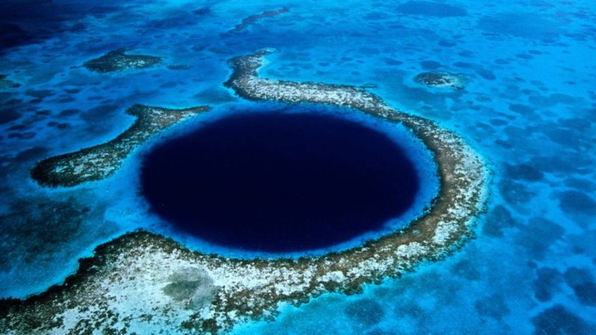 Aerial view of the Great Blue Hole, a large dark circular marine sinkhole surrounded by shallow turquoise water and coral reef.