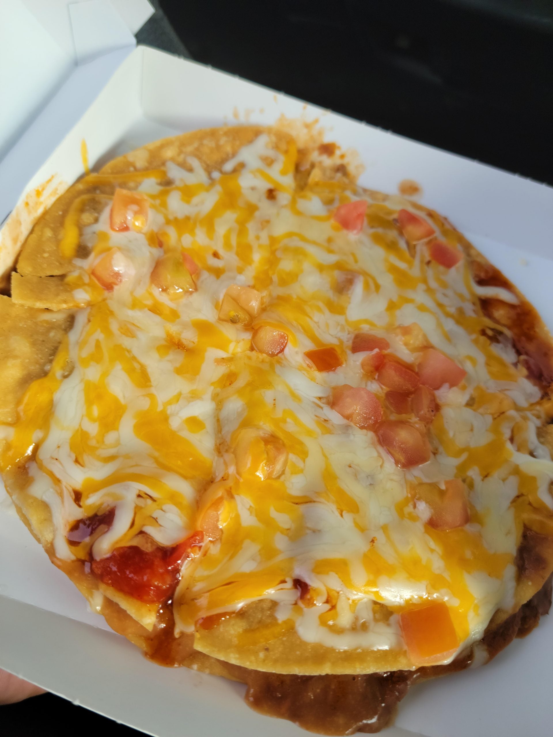 A Mexican pizza topped with melted cheese, diced tomatoes, and sauce in a cardboard takeout box.