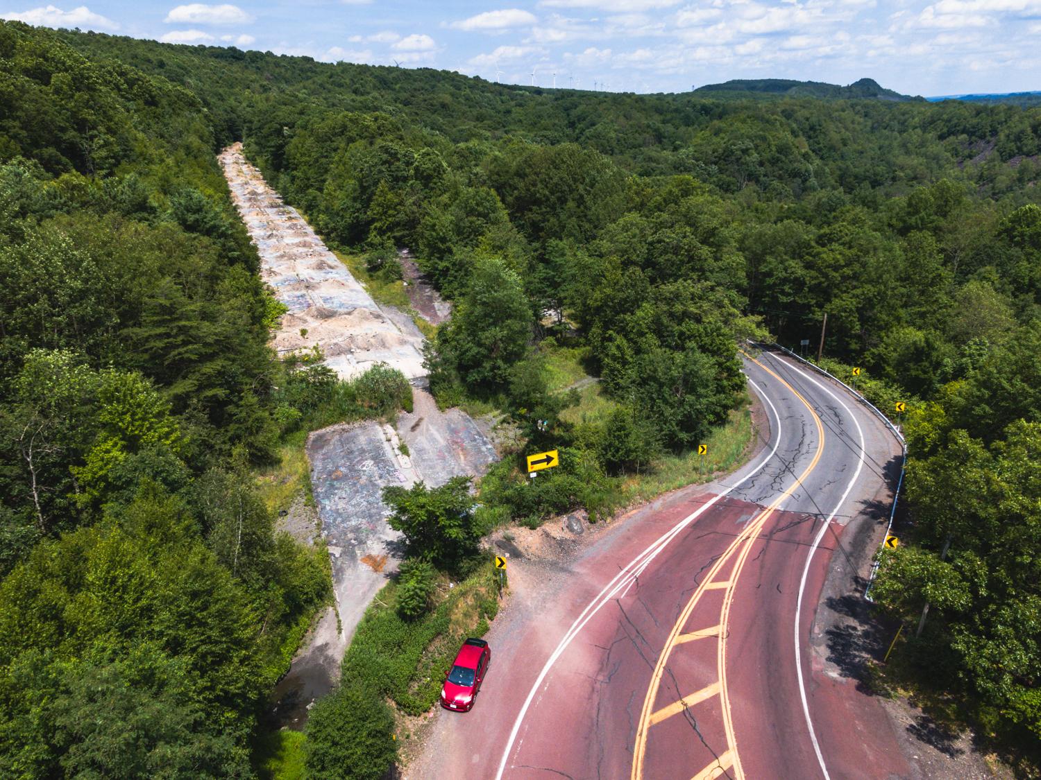 An aerial view of a red car parked on the shoulder of a curved road, next to a disused, debris-covered former roadway surrounded by dense forest.