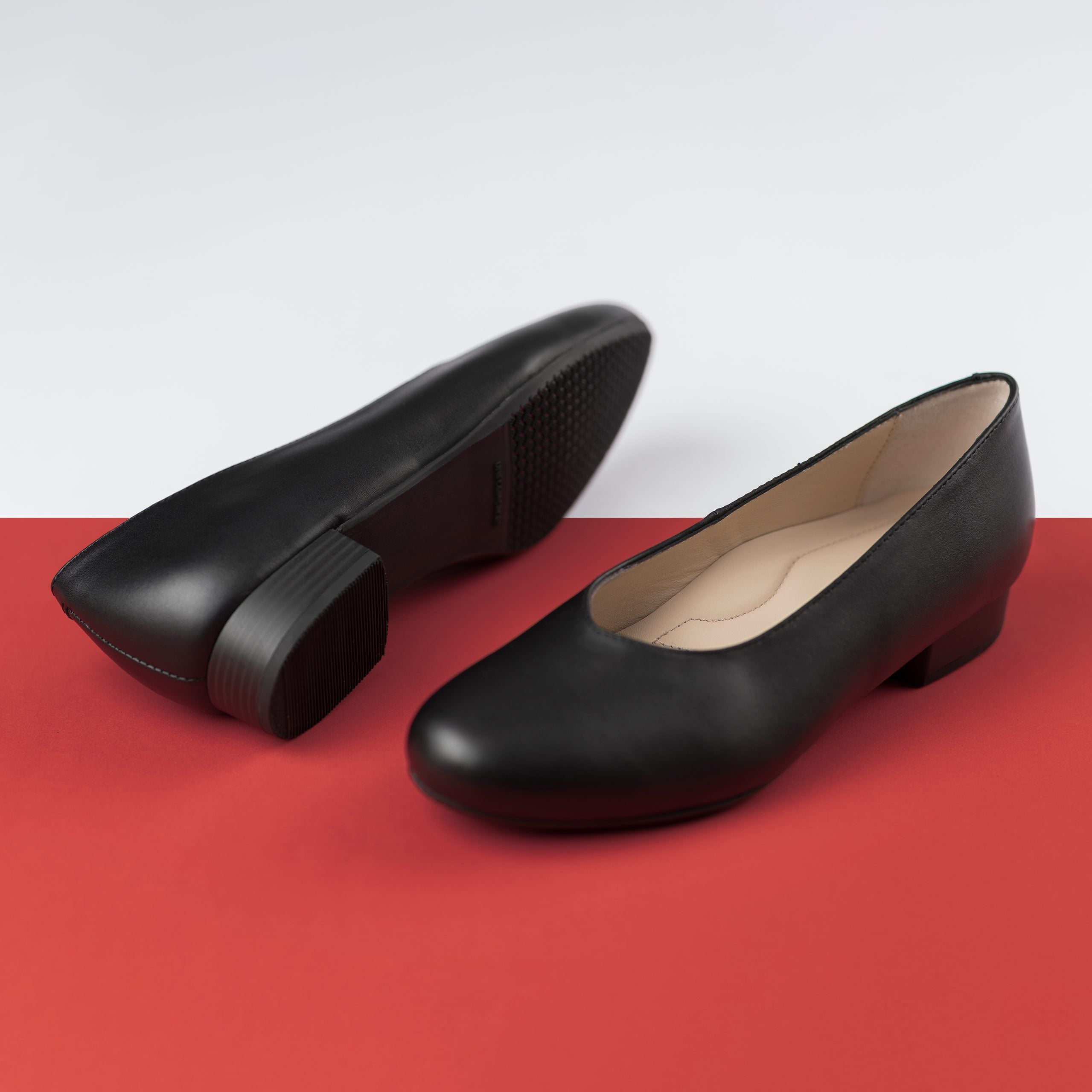 Two black round-toe flats with low heels positioned on a split white and red background.