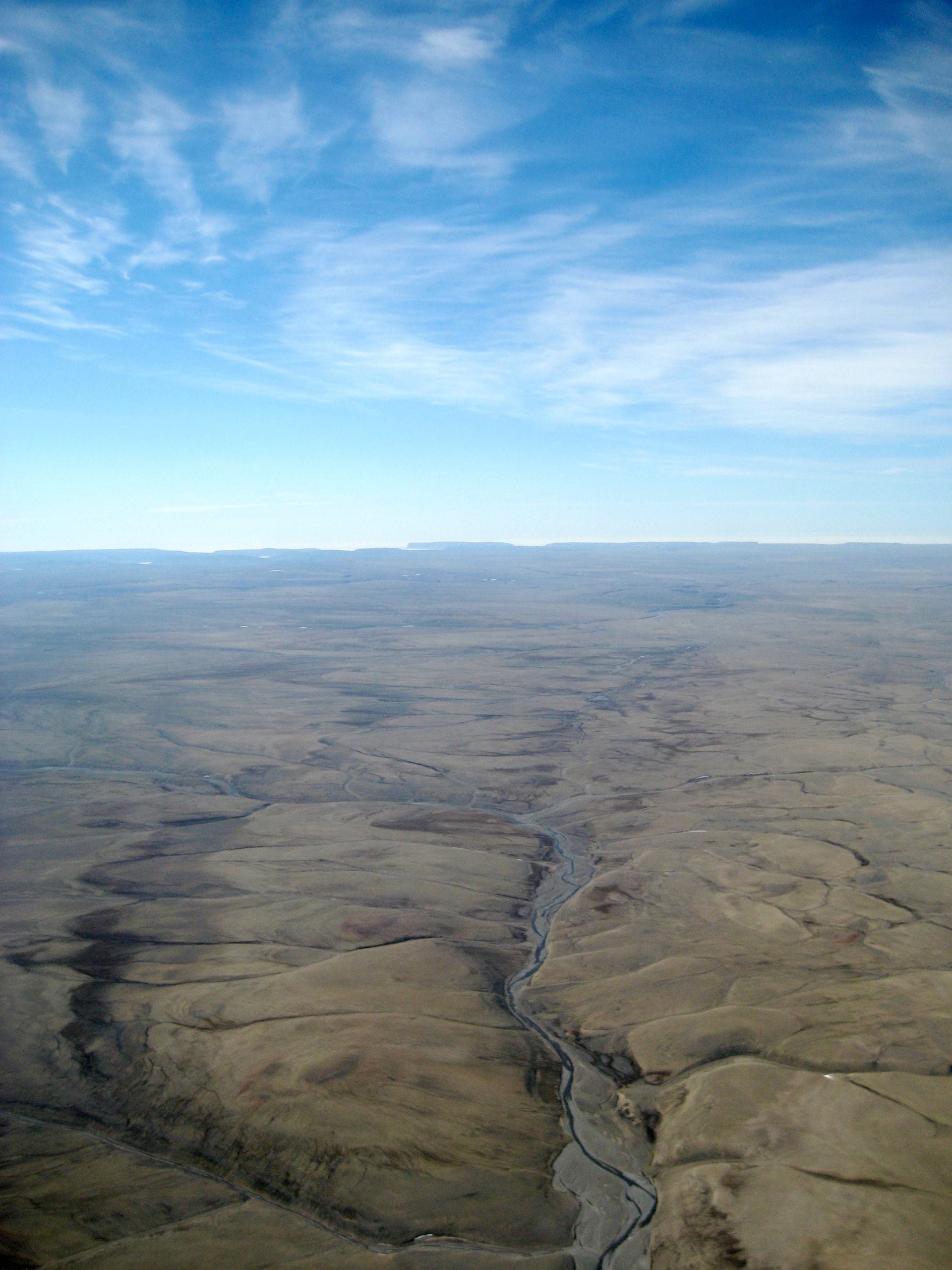 Aerial view of a vast, barren desert landscape with a single winding riverbed cutting through the terrain under a blue, cloudy sky.