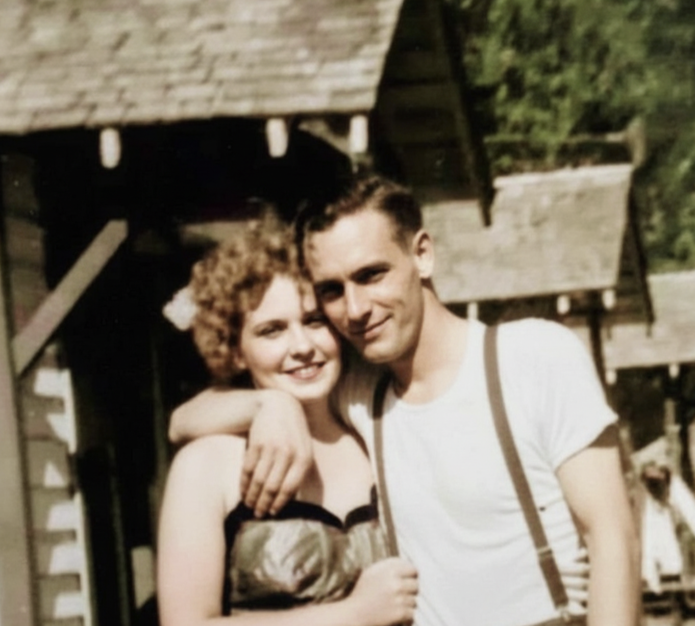 A young woman with curly hair and a man wearing suspenders pose together with their arms around each other in front of a building.