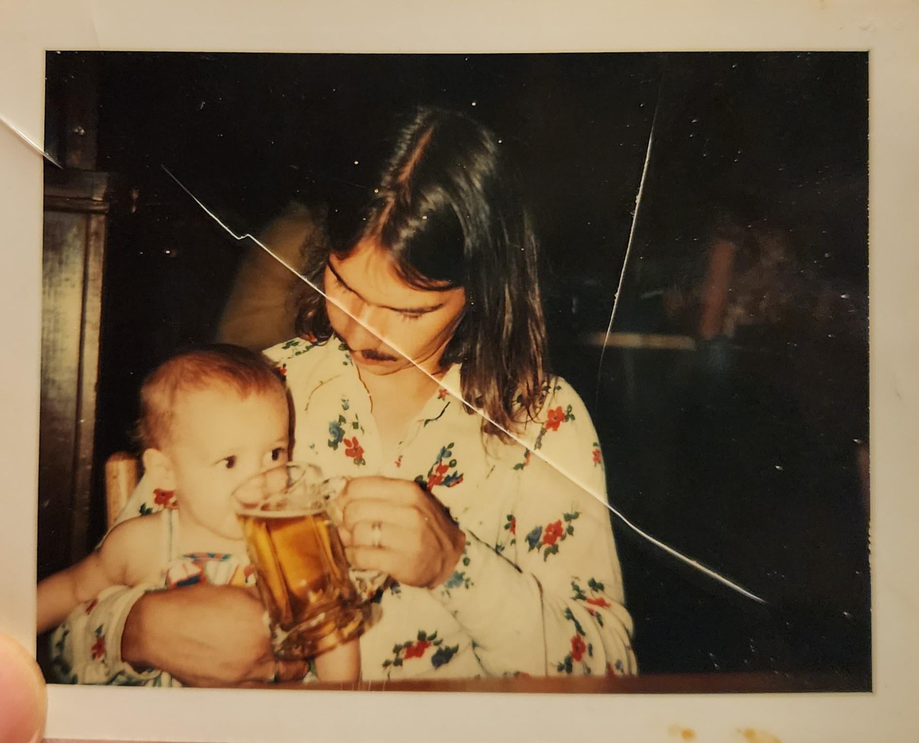 A person with long hair and a mustache holding a baby who is drinking from a glass beer mug.