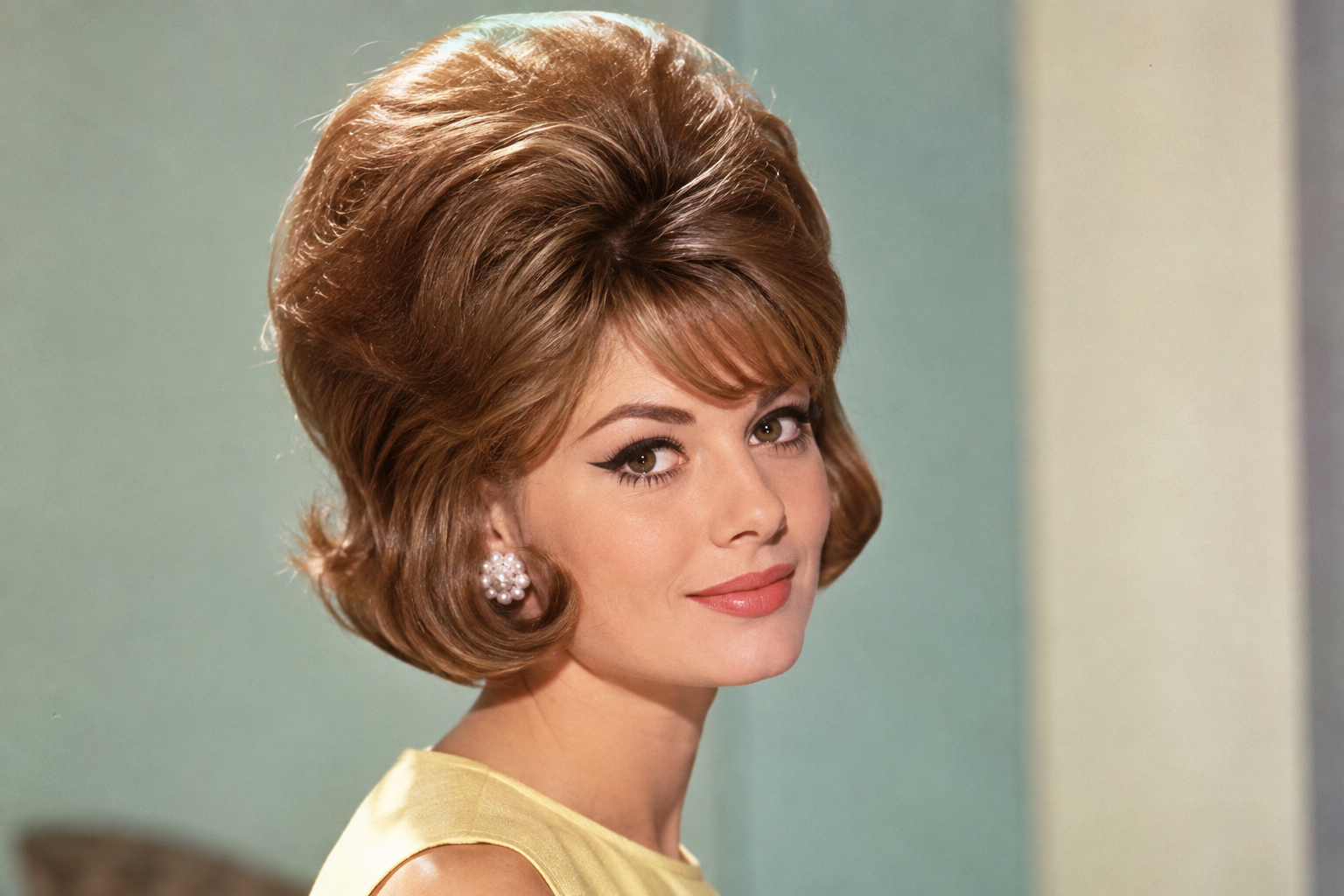 A woman with a 1960s bouffant hairstyle and winged eyeliner looks toward the camera.