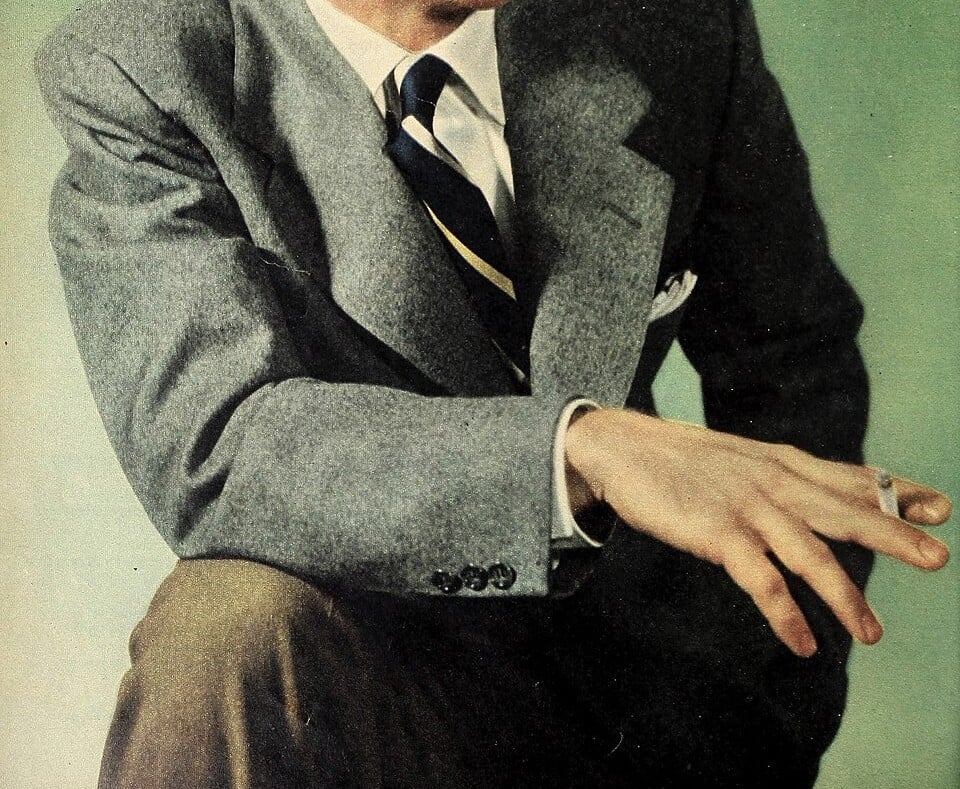 A person in a gray suit and striped tie sits with one arm resting on their knee, holding a cigarette between their fingers, against a light green background.