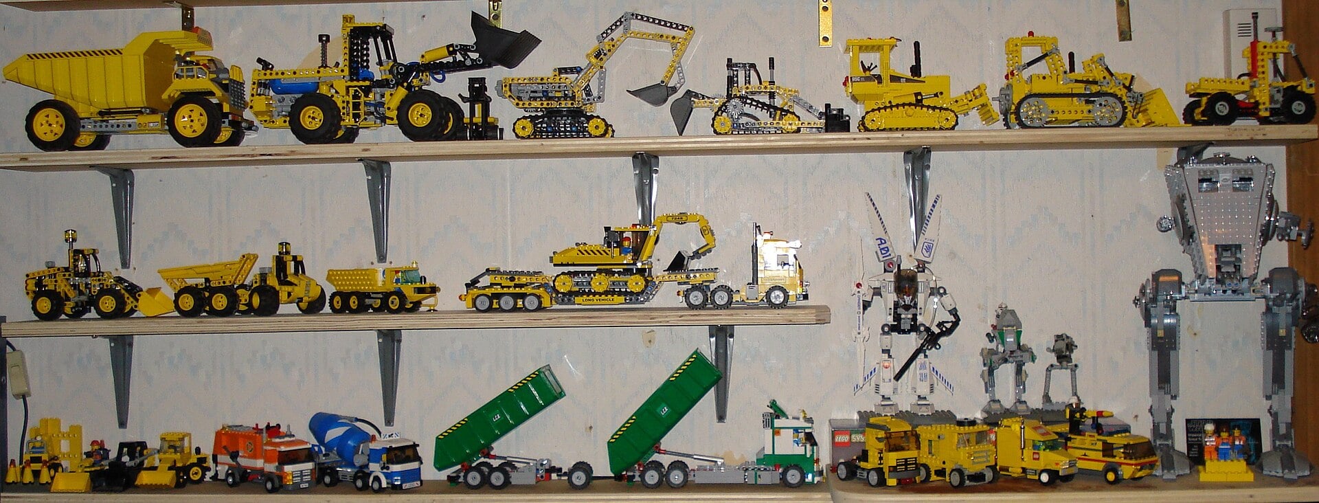 Shelves display various yellow LEGO construction vehicles, trucks, and robots, including dump trucks, bulldozers, excavators, cranes, cement mixers, and sci-fi-themed machines, all neatly arranged against a patterned wall.