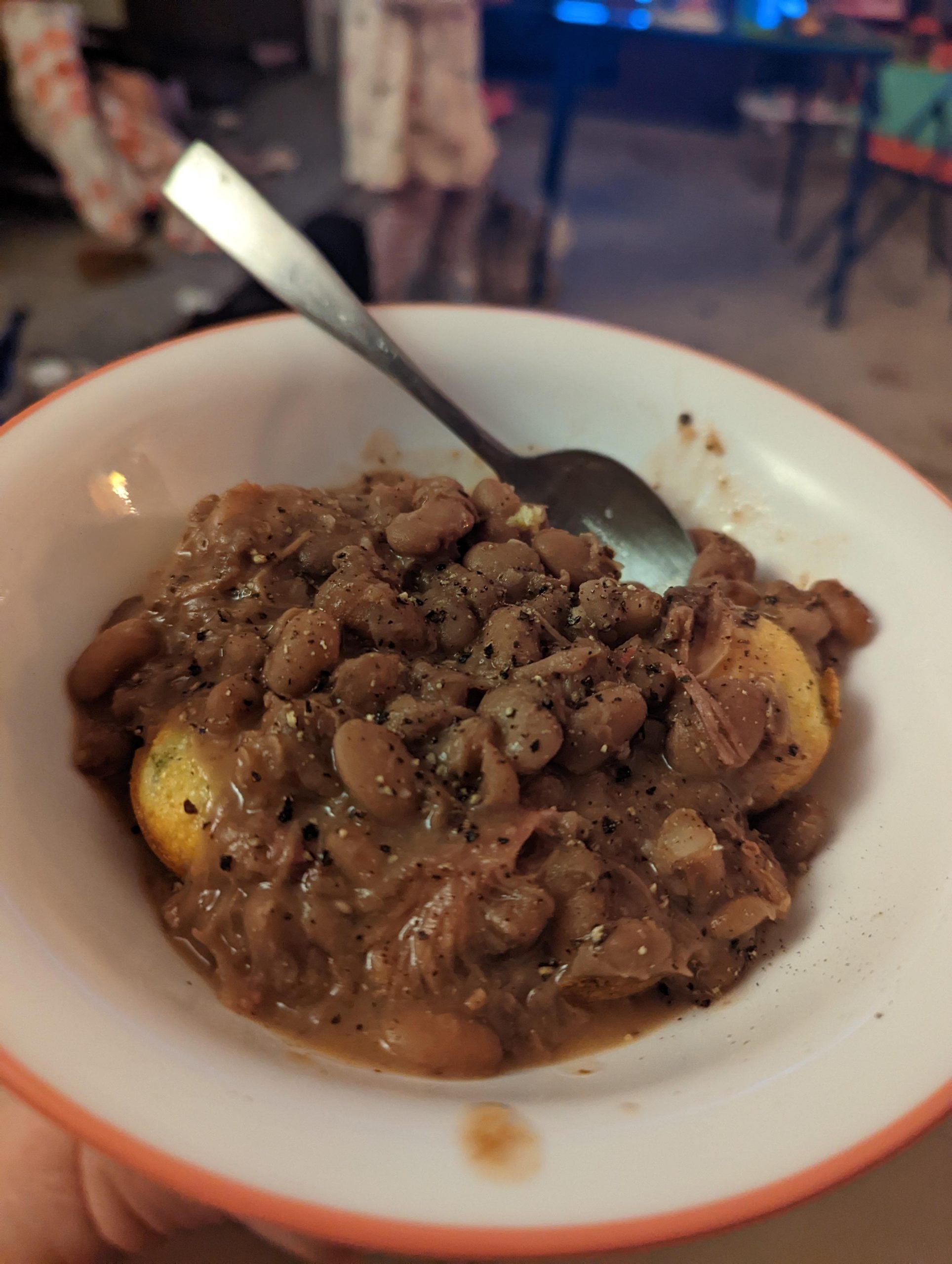 A bowl of cornbread topped with cooked beans and black pepper, with a spoon in the bowl. The background is blurry, showing a room with scattered furniture and objects.