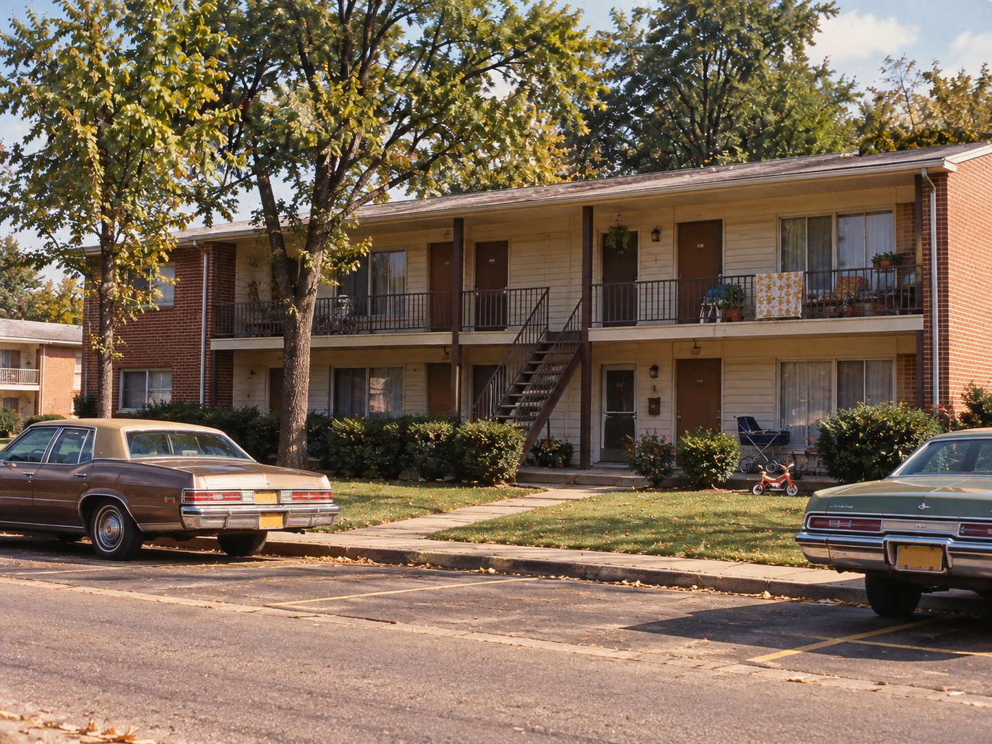 A two-story apartment building with exterior walkways and staircases, flanked by parked cars.