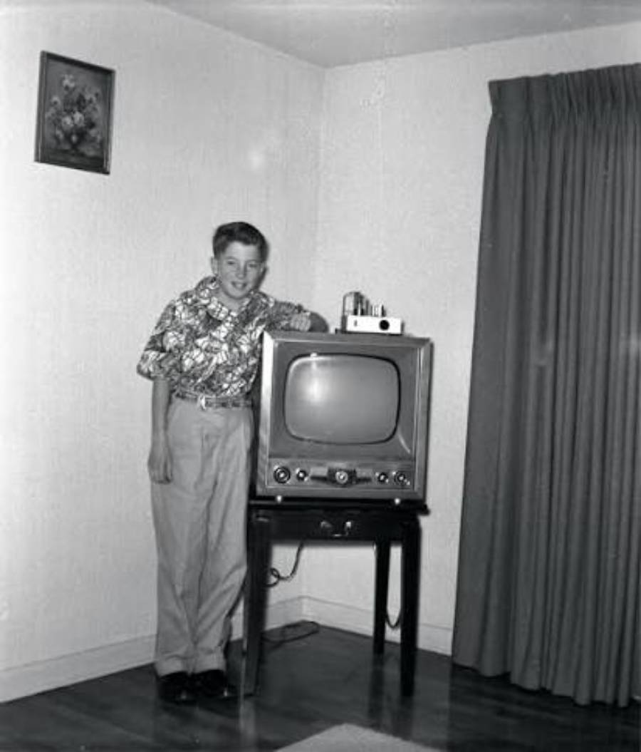 A young boy standing next to a vintage television on a stand in a living room.