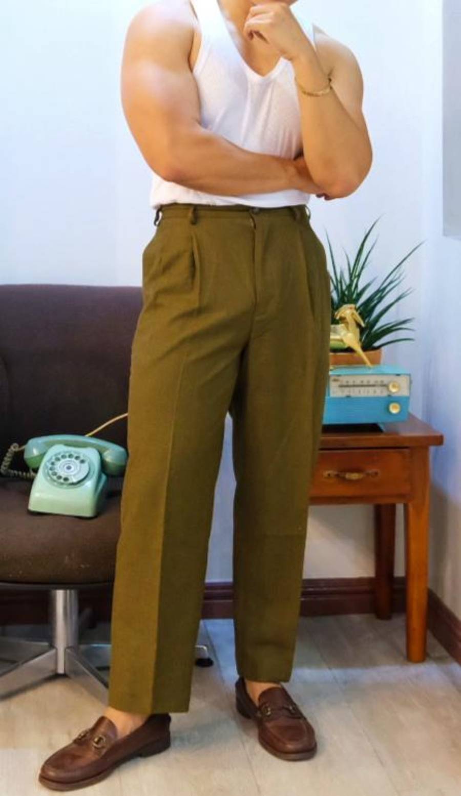 A person in a white tank top, olive green high-waisted pants, and brown loafers stands indoors by a chair, a small table with a green rotary phone, a radio, and a plant. Their face is not visible.