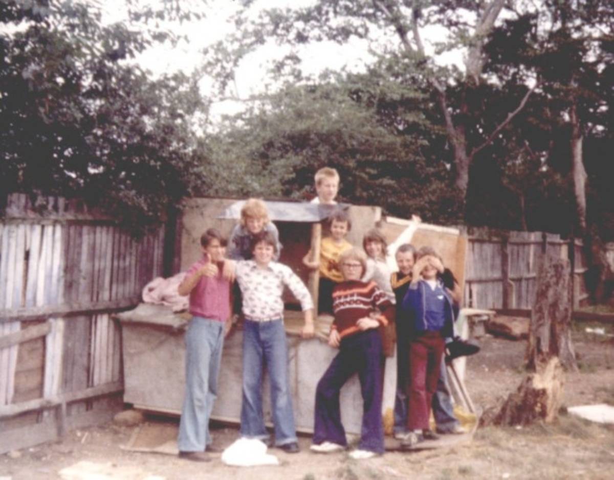 A group of seven children standing around and inside a homemade wooden play structure in a backyard.