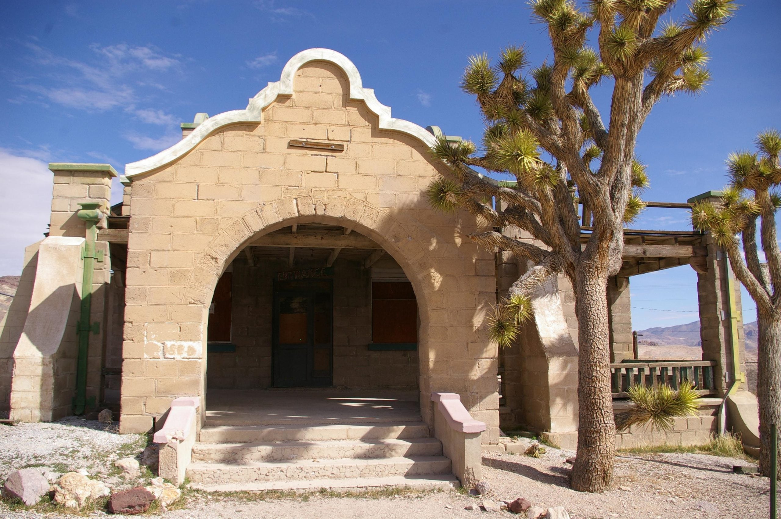 An old stone building with a large arched entryway and an “ENTRANCE” sign above the door, flanked by two Joshua trees.