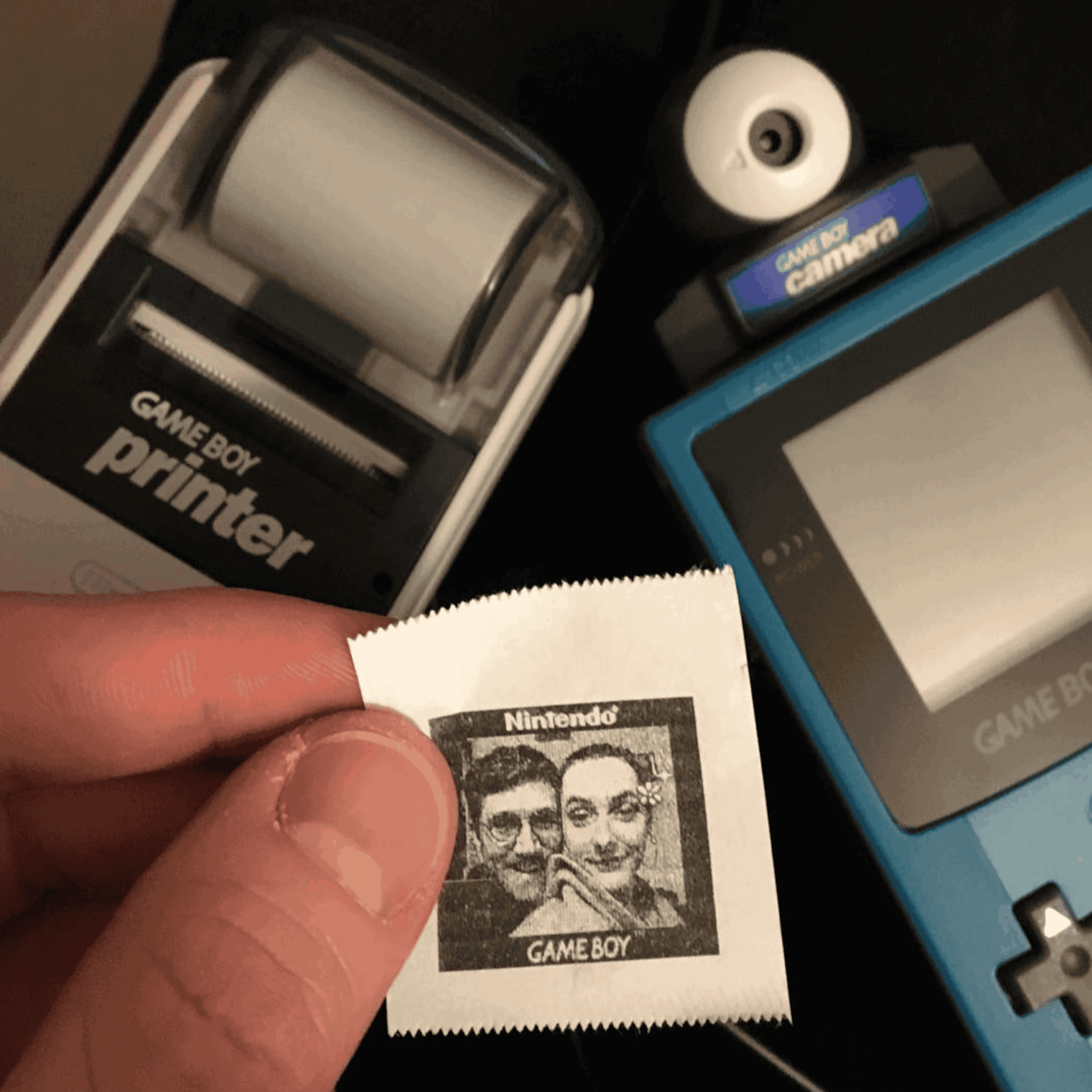 A person holds a small, black-and-white thermal print of a portrait from a Game Boy Camera, positioned between a Game Boy Printer and a blue Game Boy console with a camera attachment.