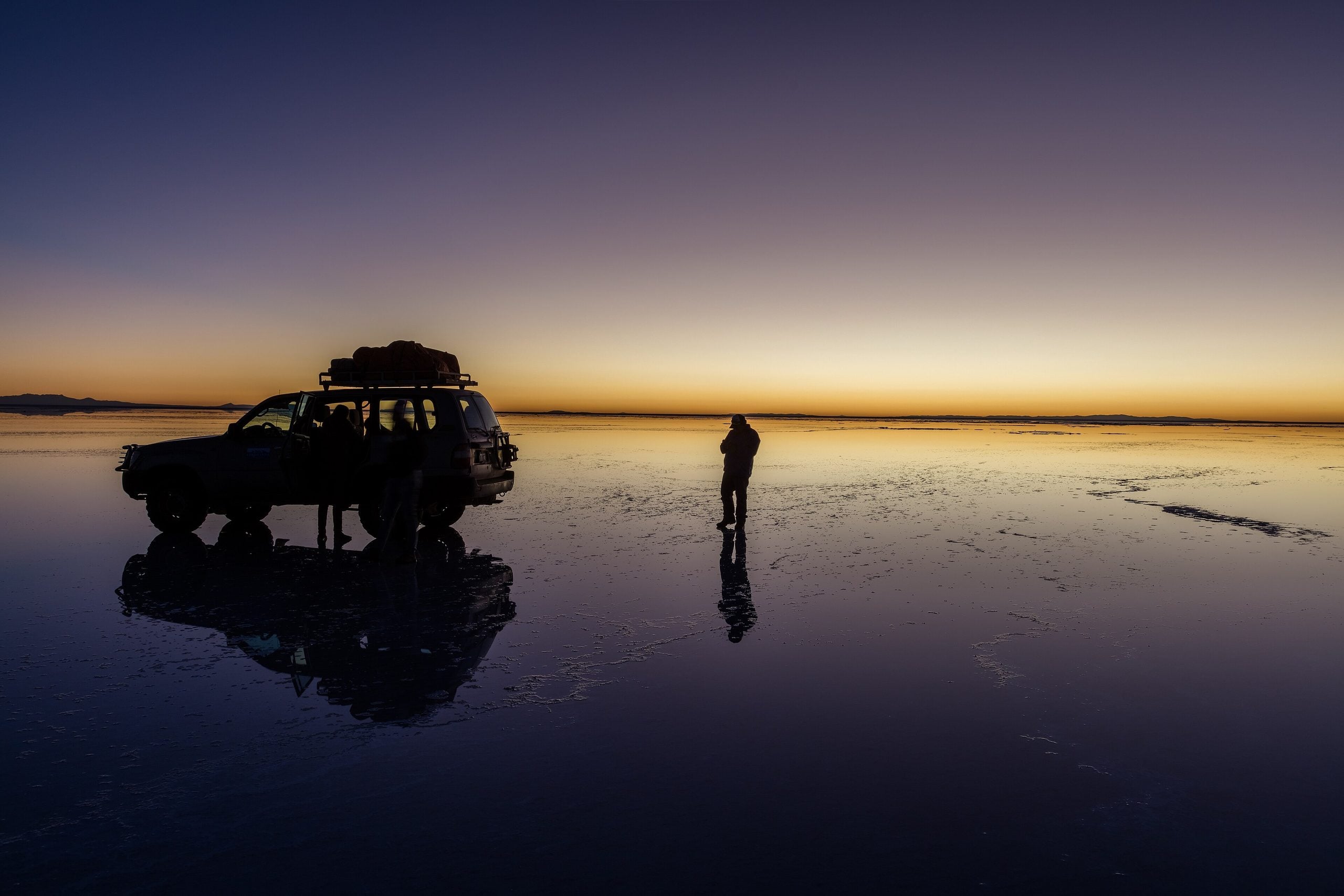A person stands next to an SUV parked on the reflective surface of the Salar de Uyuni salt flat at sunset.