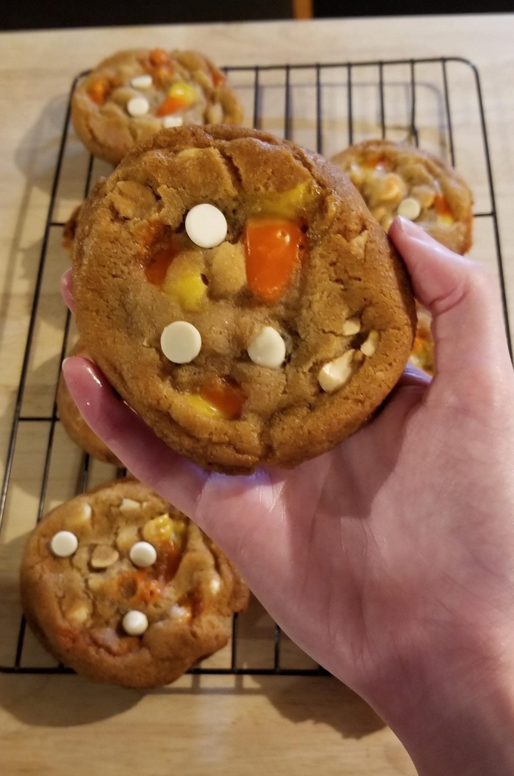 A hand holds a large cookie topped with candy corn and white chocolate chips, with more cookies resting on a wire cooling rack in the background.