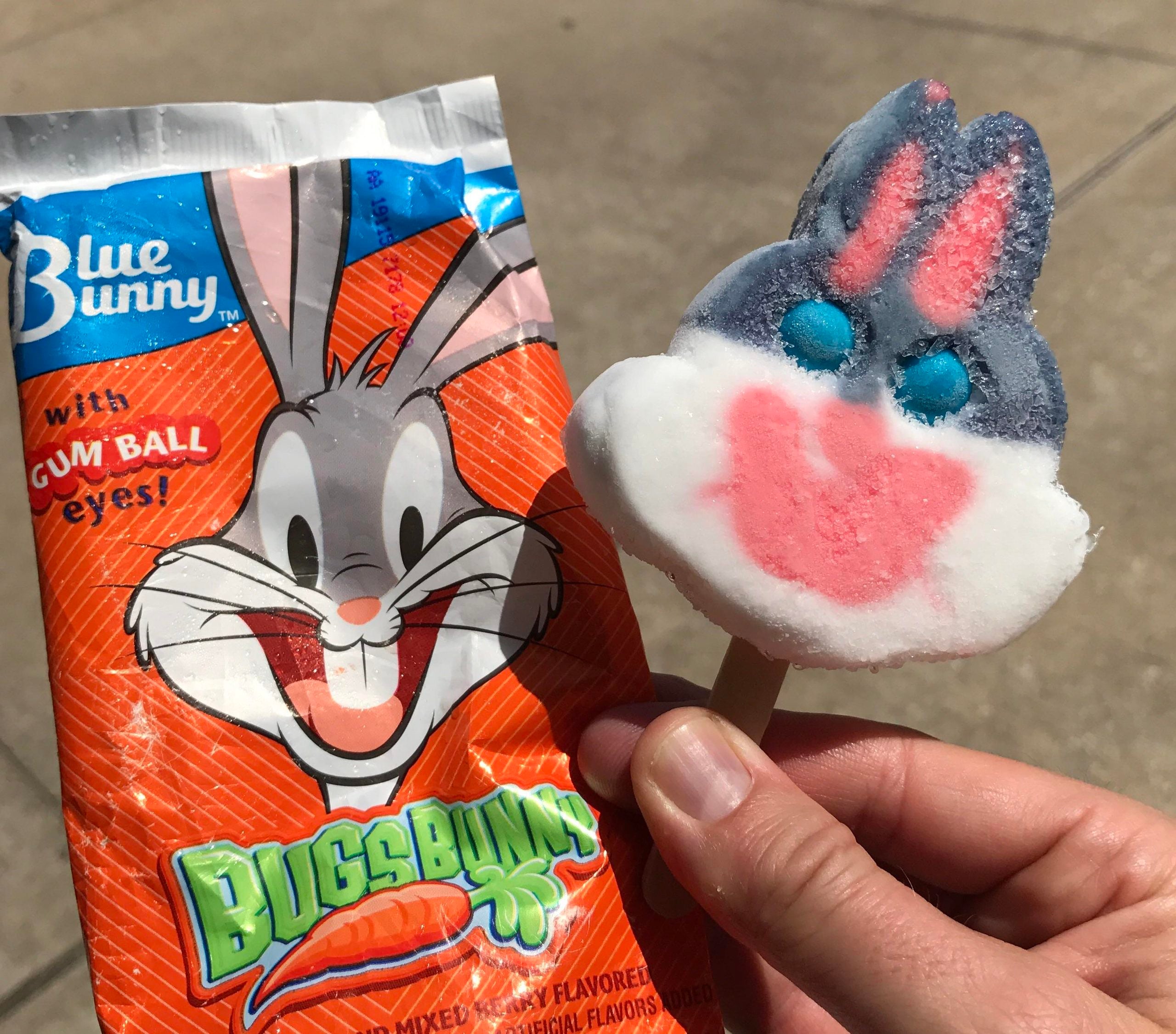 A hand holds a Bugs Bunny character popsicle next to its bright orange and blue Blue Bunny brand wrapper.