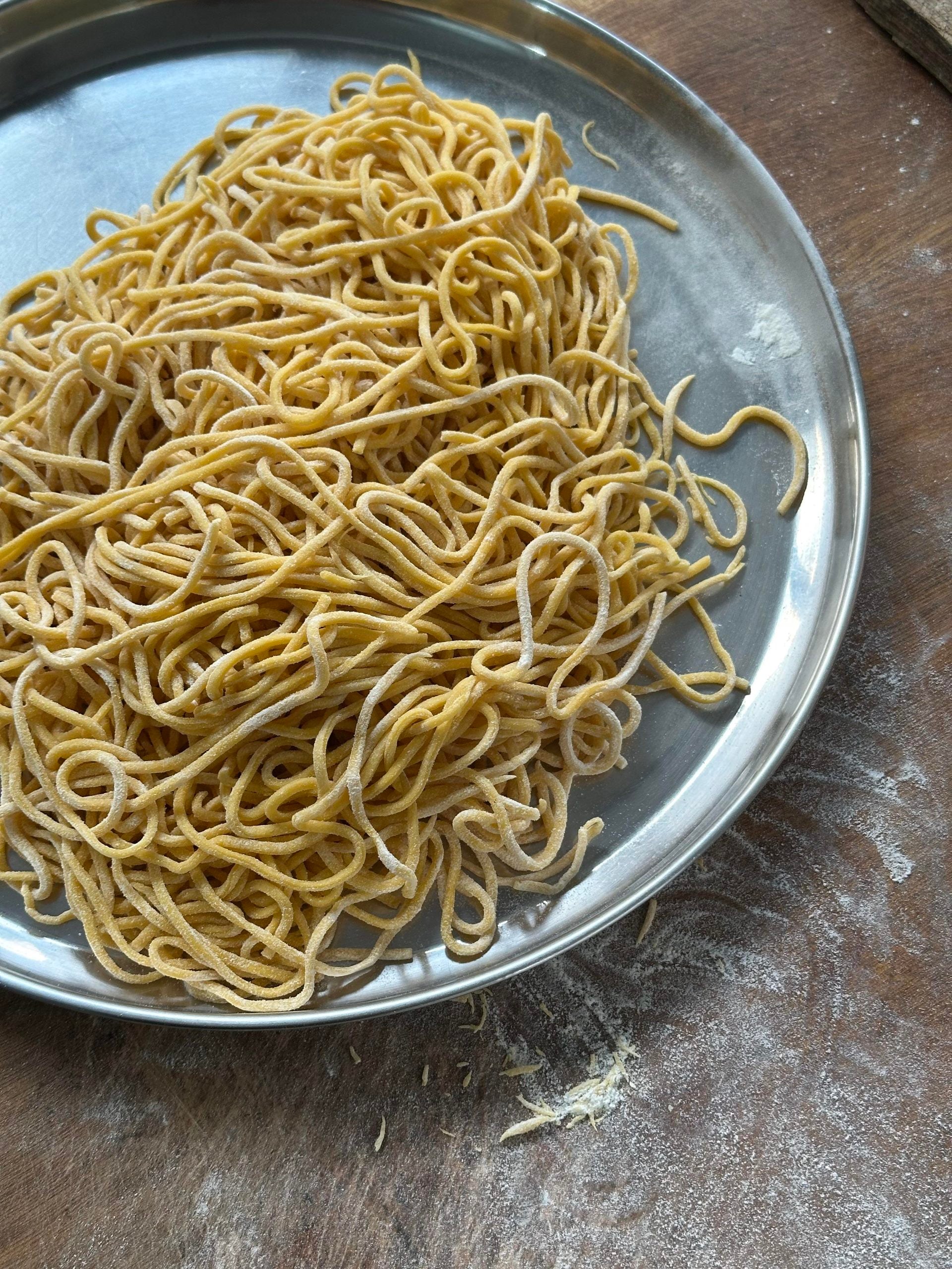 Fresh, uncooked yellow noodles are piled on a round metal plate, resting on a wooden surface dusted lightly with flour.