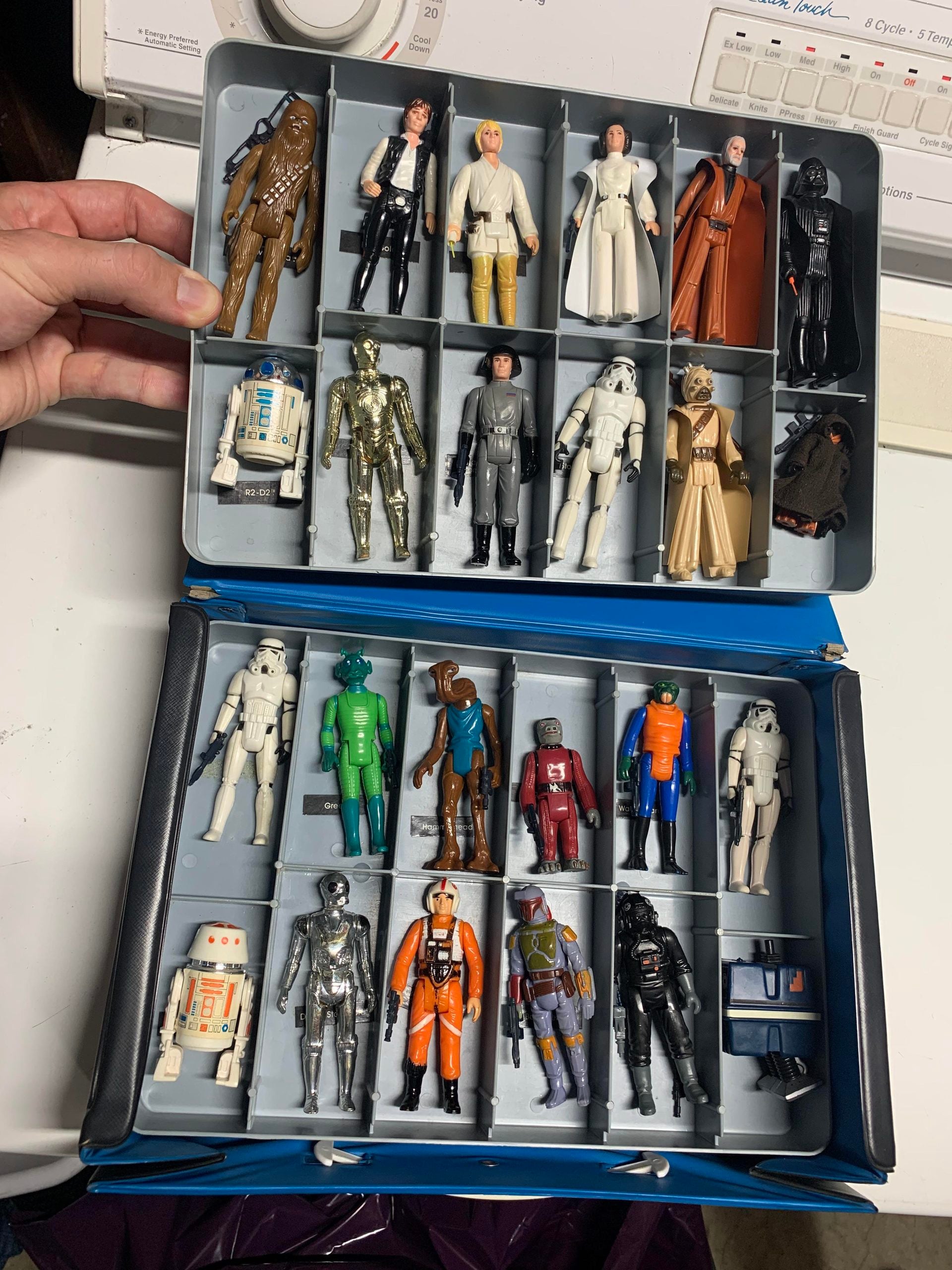 Open case with vintage Star Wars action figures arranged in compartments, including characters like Chewbacca, Luke, Leia, R2-D2, and Darth Vader. A hand holds the case, and the background shows a washing machine.
