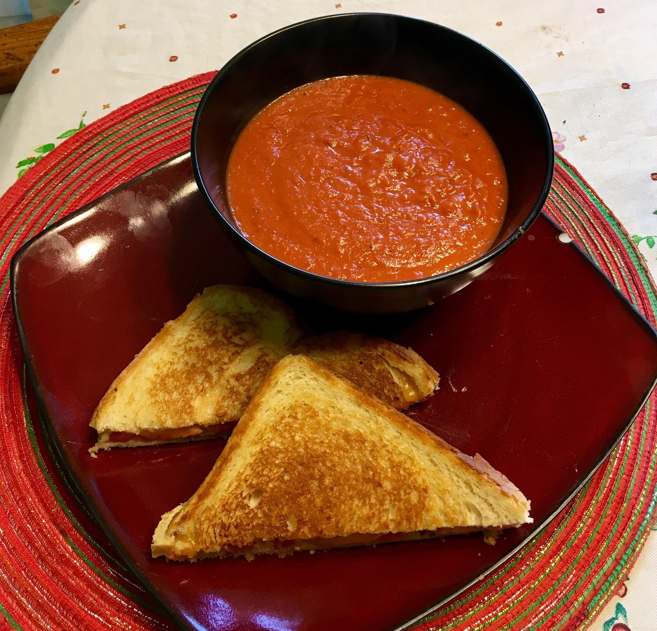 A bowl of tomato soup sits on a red plate next to two grilled cheese sandwich halves, all placed on a colorful placemat.