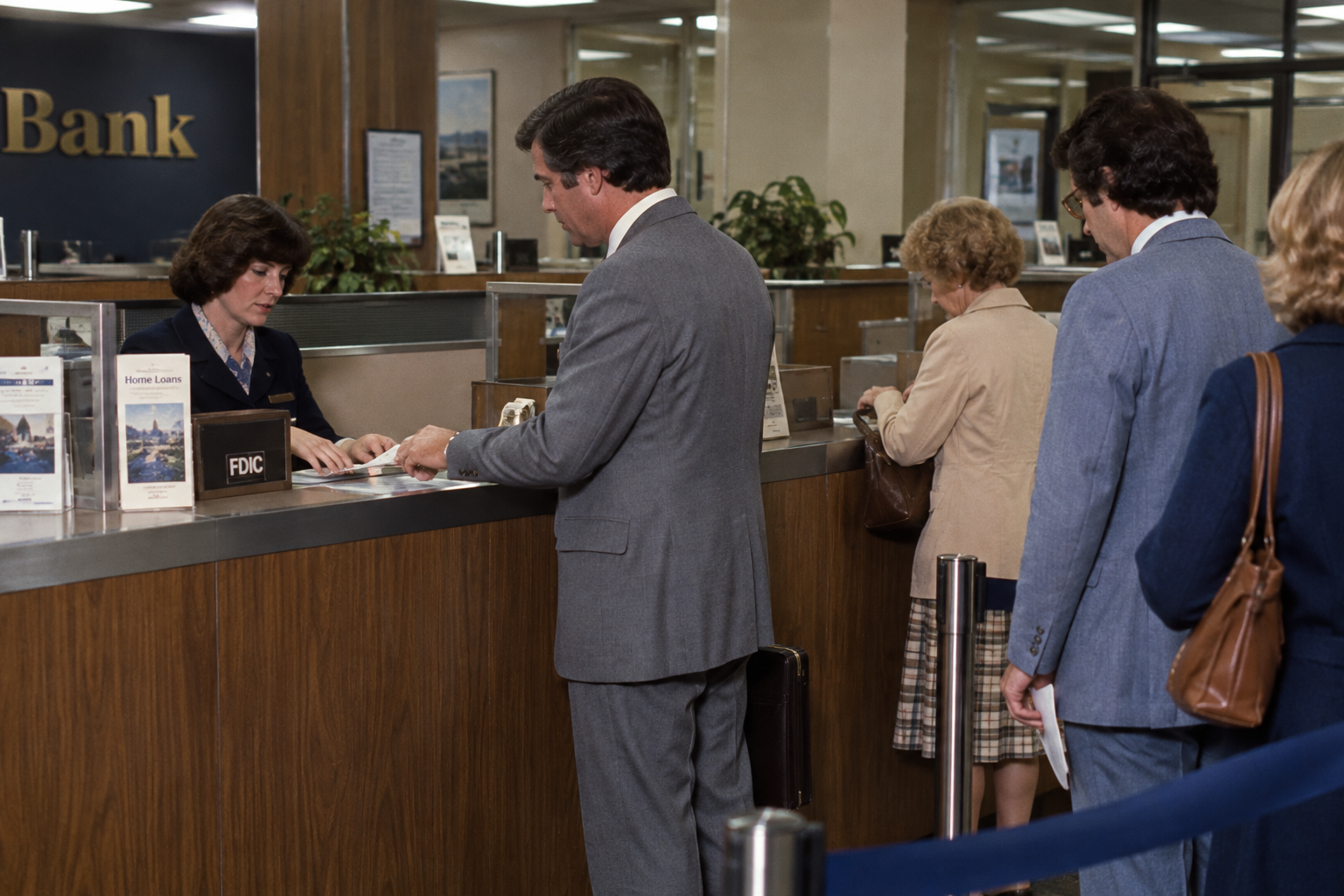 A bank teller assists a customer at a counter while others wait in line.