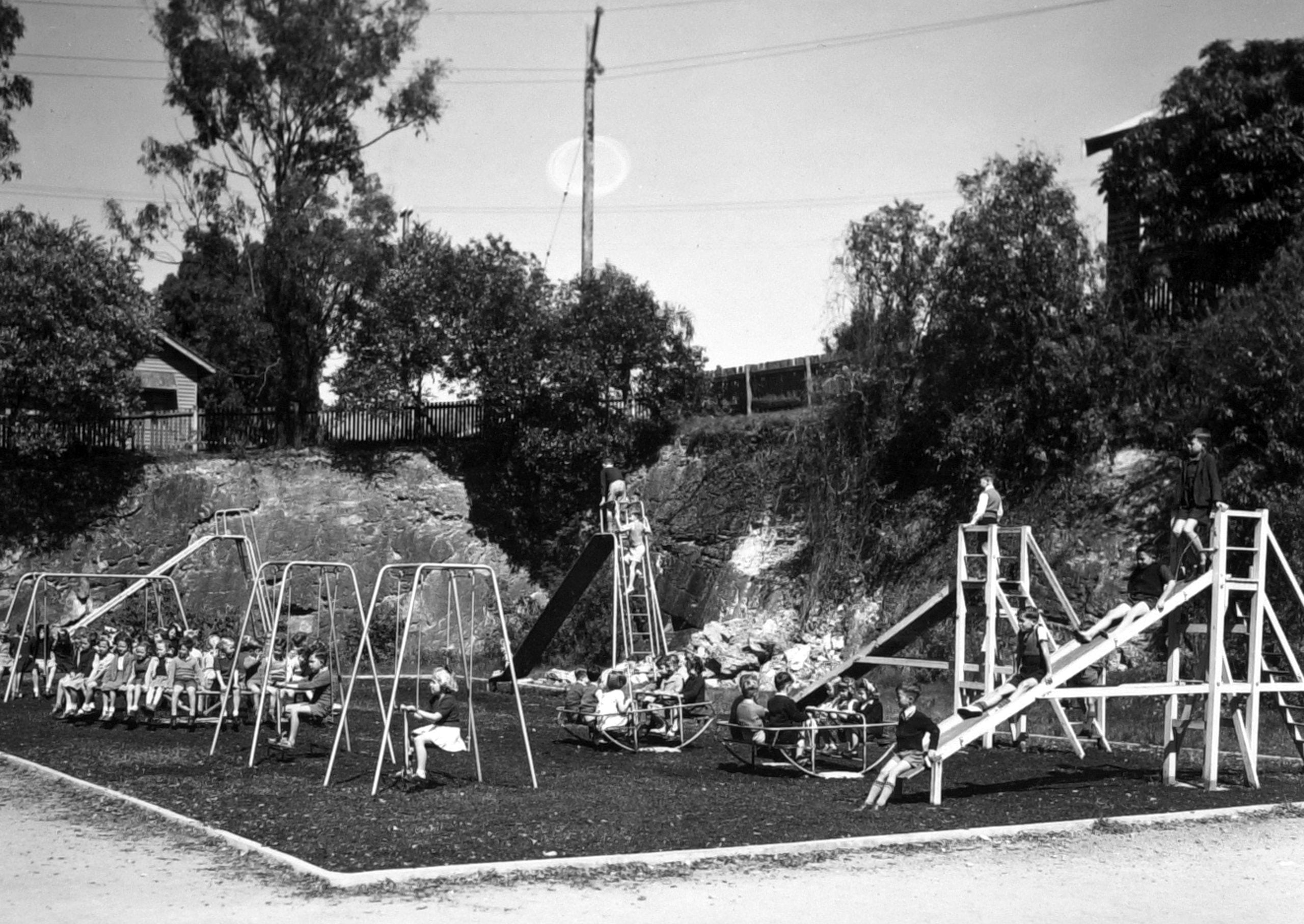 Children playing on swings, a slide, and a merry-go-round in a playground set against a rock cliff.